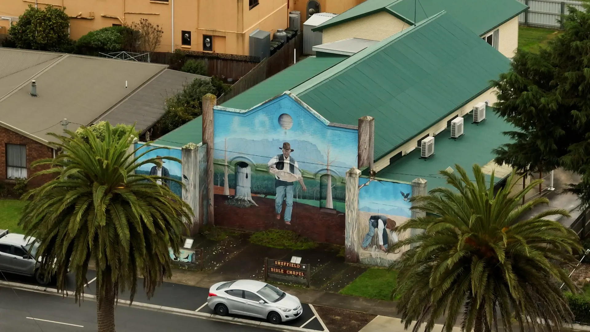 An elevated view of a green-roofed building with a large painted mural on it's street-facing facade of a man in a hat and vest.