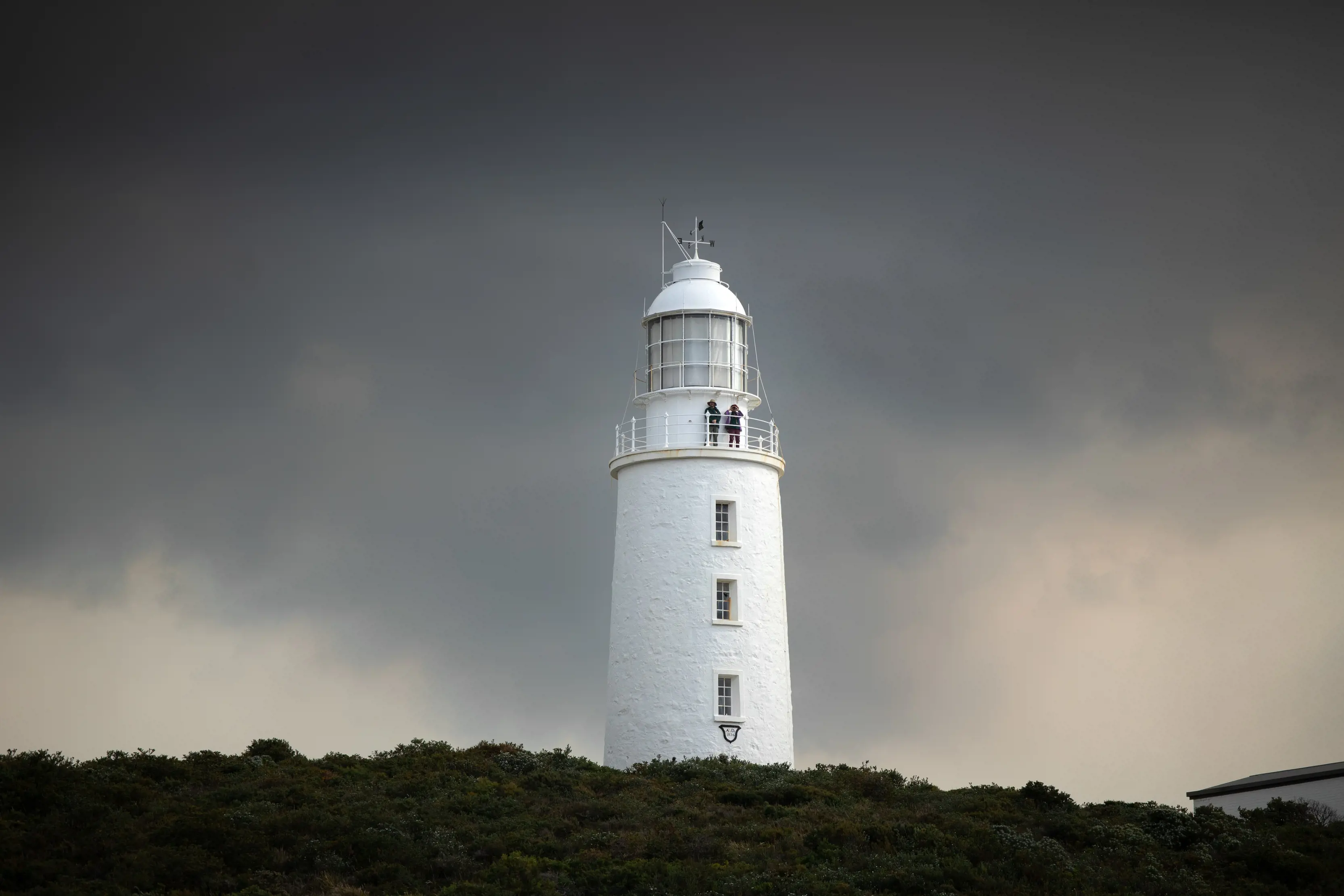 Exterior of two people standing on the lookout at Cape Bruny Lighthouse, with grey clouds in the sky.