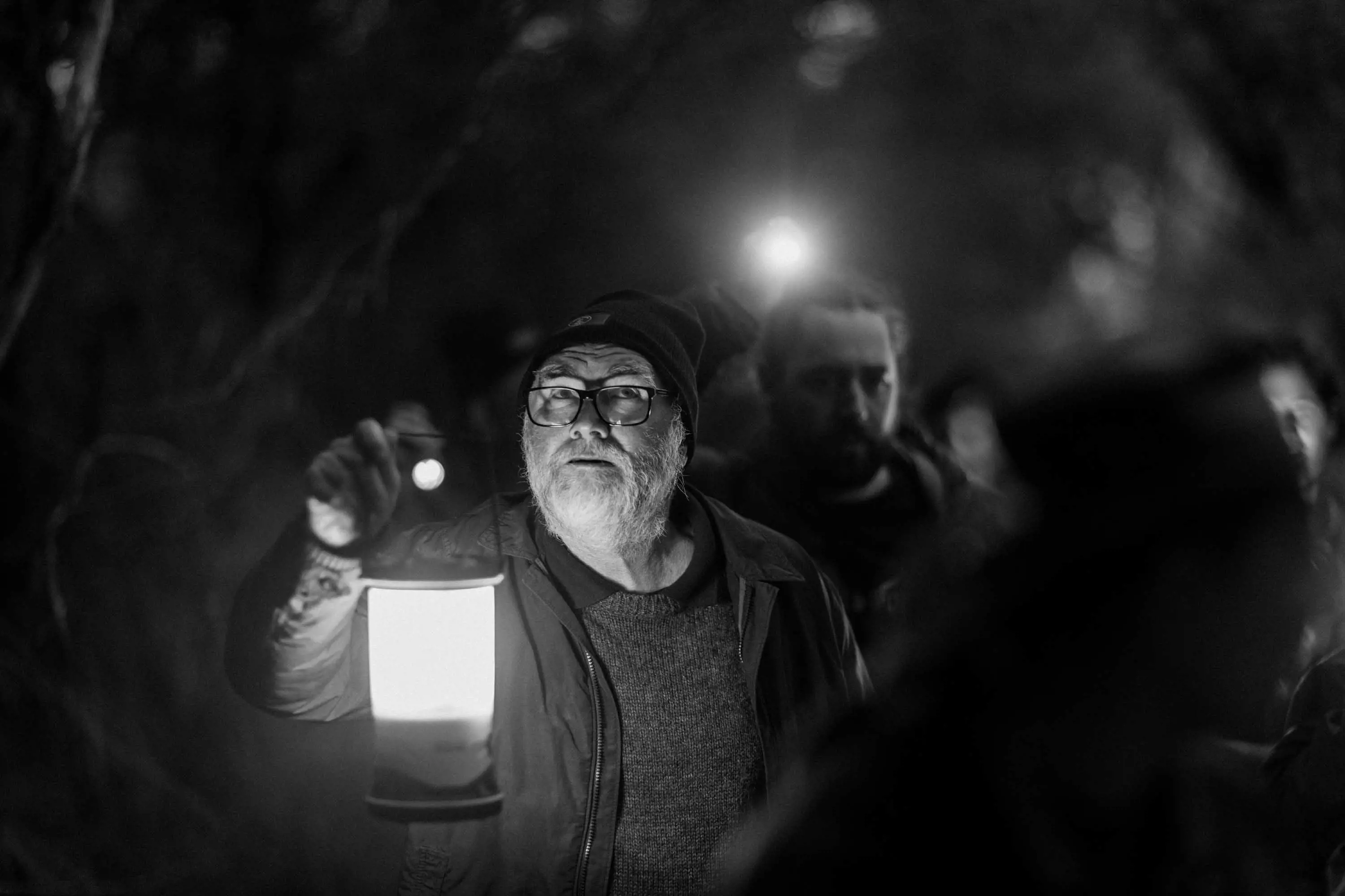 A senior man with a beard, wearing a beanie holds a lantern and guides a group of people through darkness.