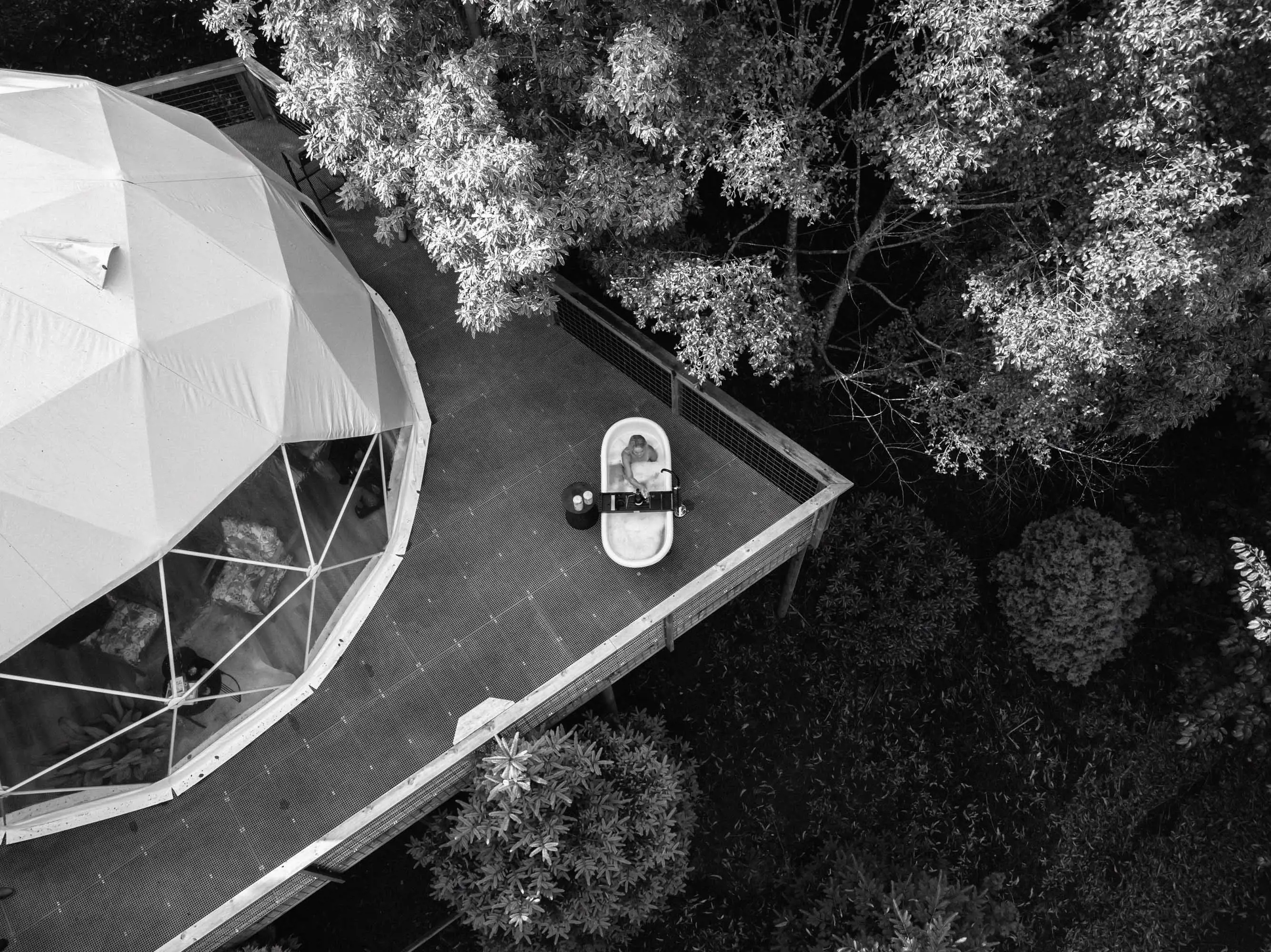 An aerial view of a large icosahedron-shaped tent with large, triangular window panels sits on a platform in the forest. A woman sits in a bath in one corner of the platform.
