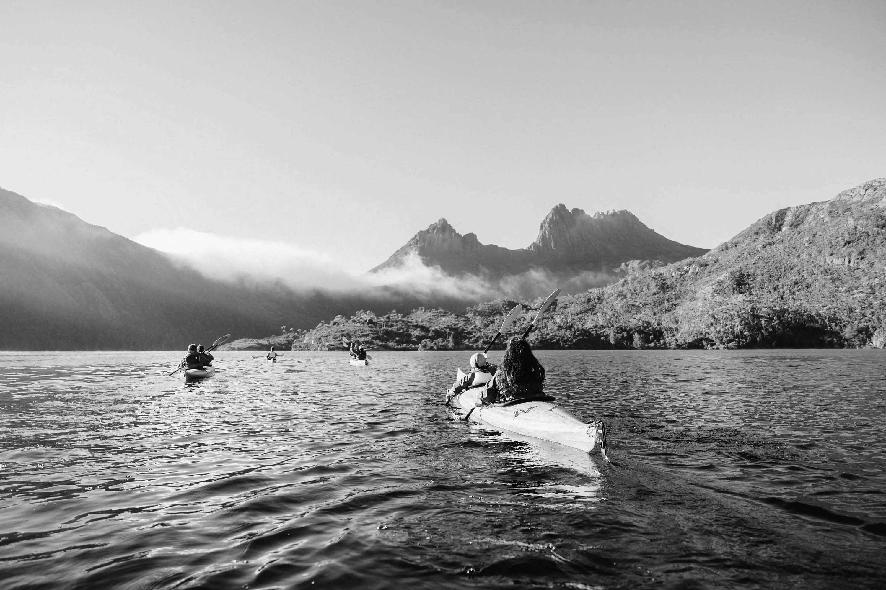 Four canoes float across a lake in front of the tall, dramatic peaks of Cradle Mountain on a clear day.