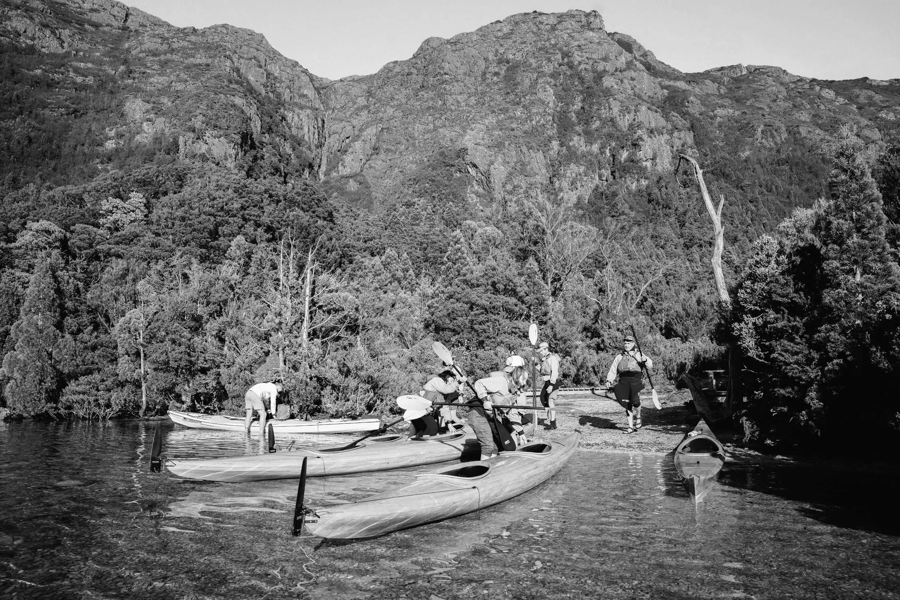 A group of people pull three canoes sit in shallow water at the edge of water with tall peaks in the background.