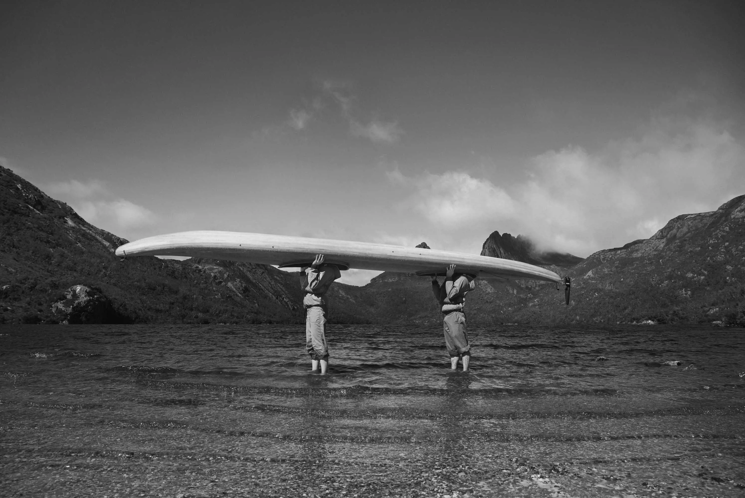 Two people hold a large canoe over their head and stand in the shallow water at the edge of a large lake in front of mountains.