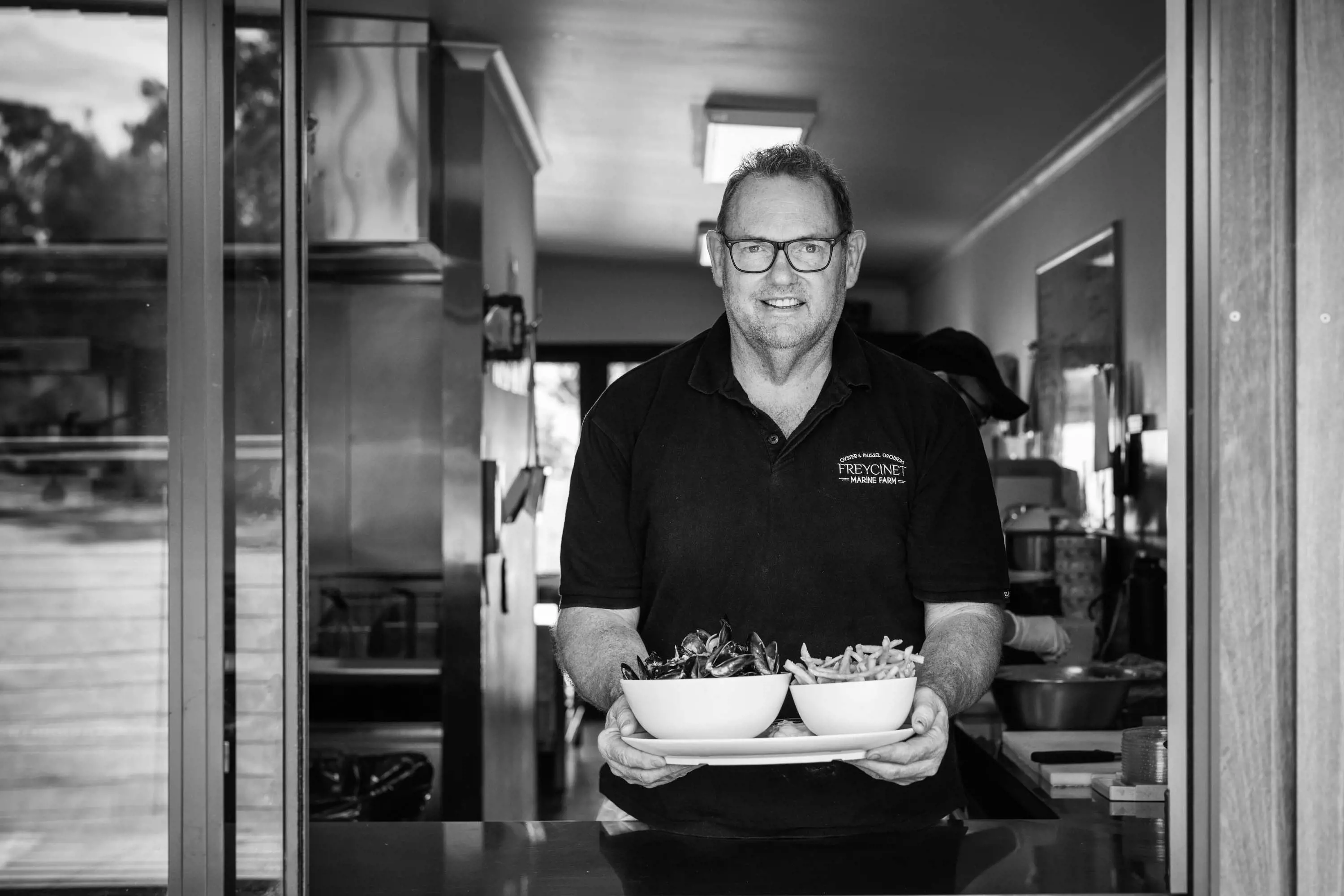 A man wearing glasses and a polo shirt stands in a doorway holding freshly prepared mussels and fried chips.