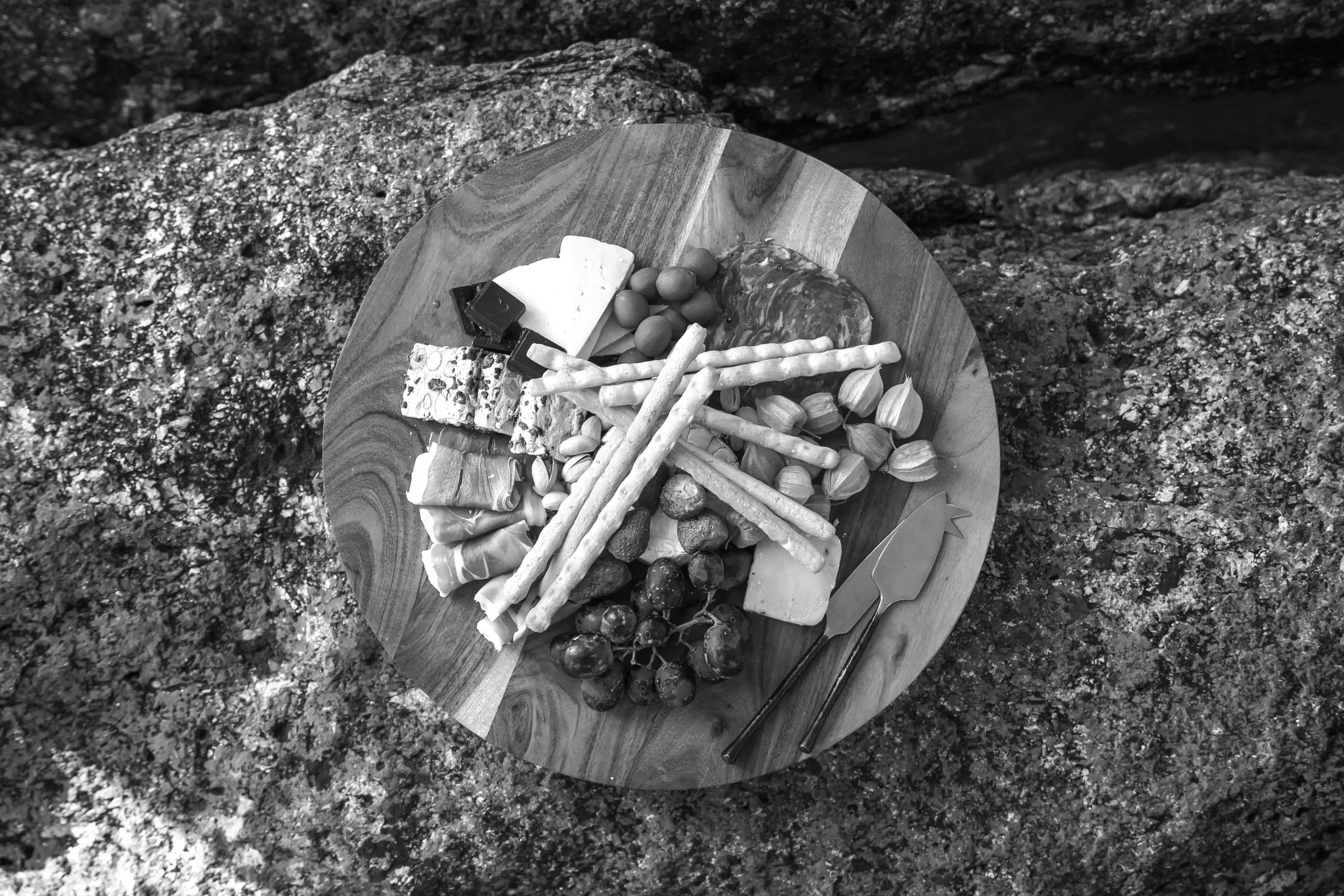 A wooden platter of meats, cheese, dates and bread sticks placed on rocks.