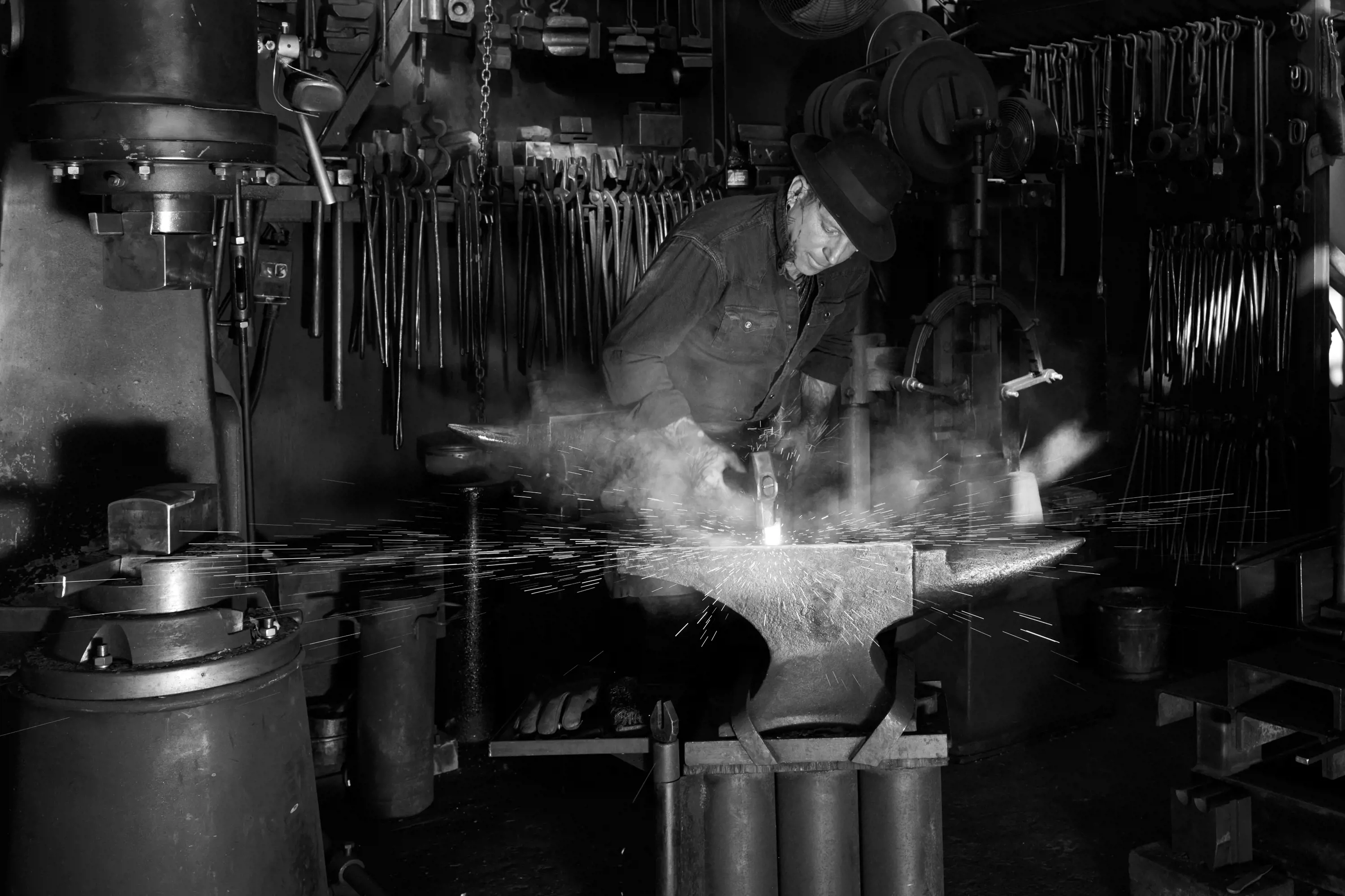 A man stands in the shadows of a metal workshop and hits hot steel on an anvil with a hammer. Sparks and smoke fill the air.