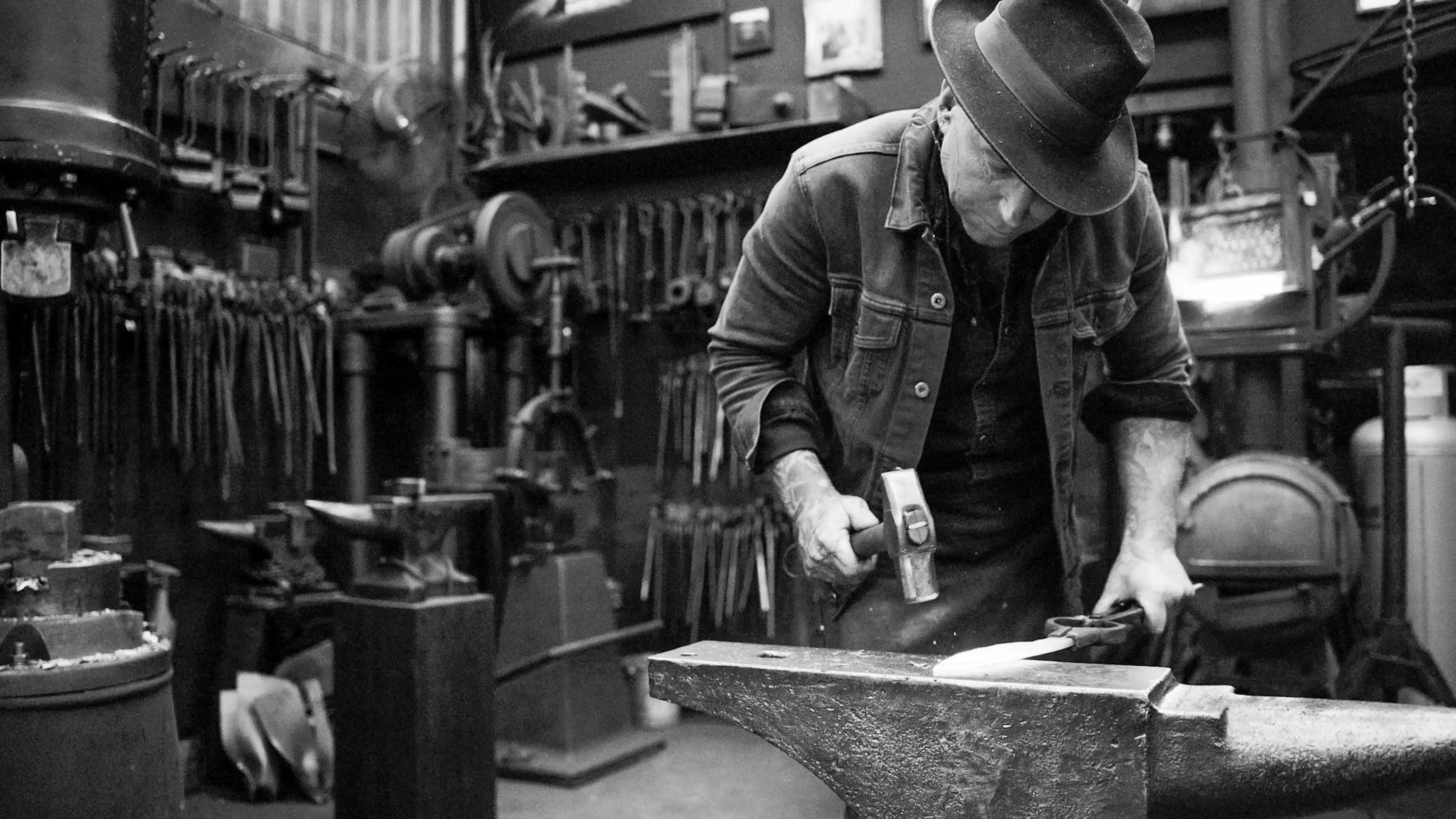 A man wearing a hat works at a piece of white-hot steal on an anvil with a metal hammer.
