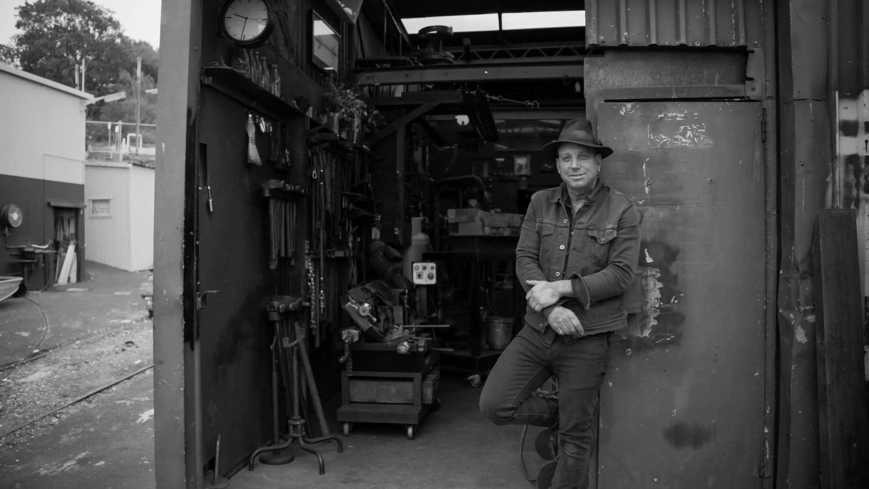 A man wearing a hat, denim jacket, black jeans and boats leans against the open door of a metal workshop.