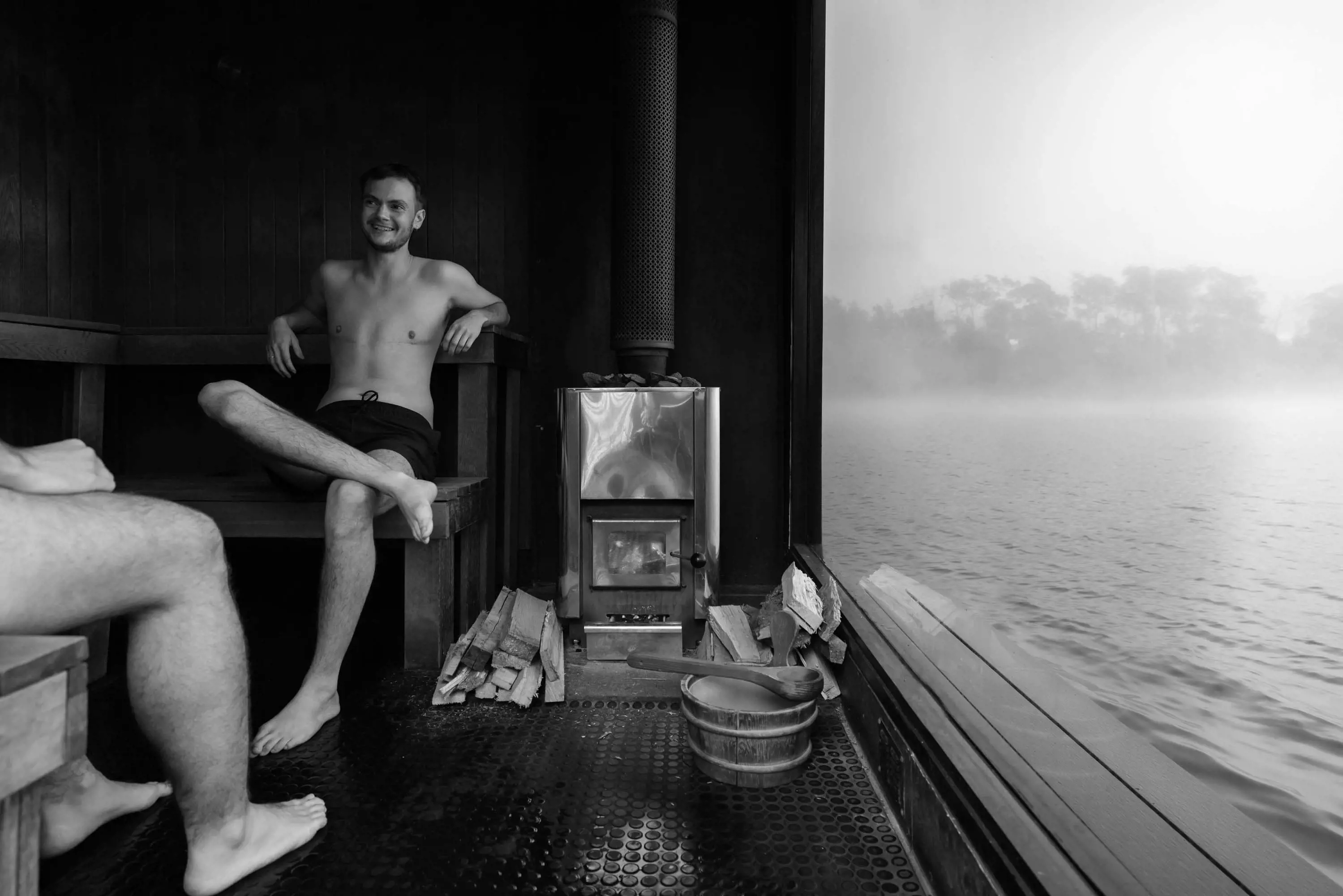 A couple sit inside a large sauna room with a floor-to-ceiling window looking out over a misty lake.
