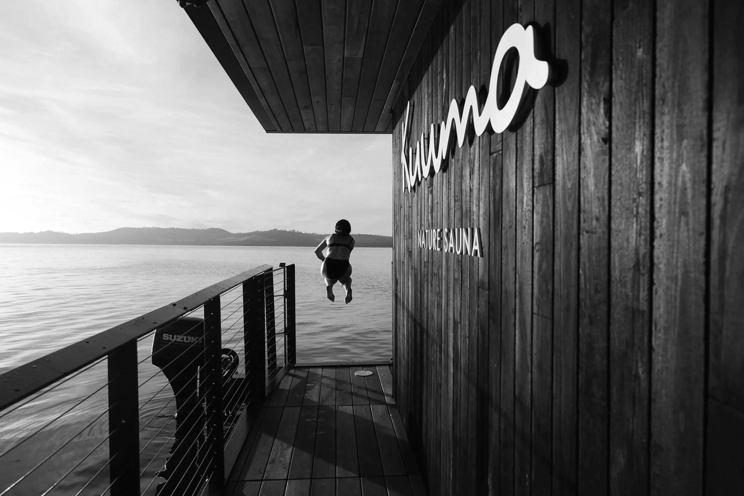 A woman jumps from the end of a patio attached to a floating sauna into water.