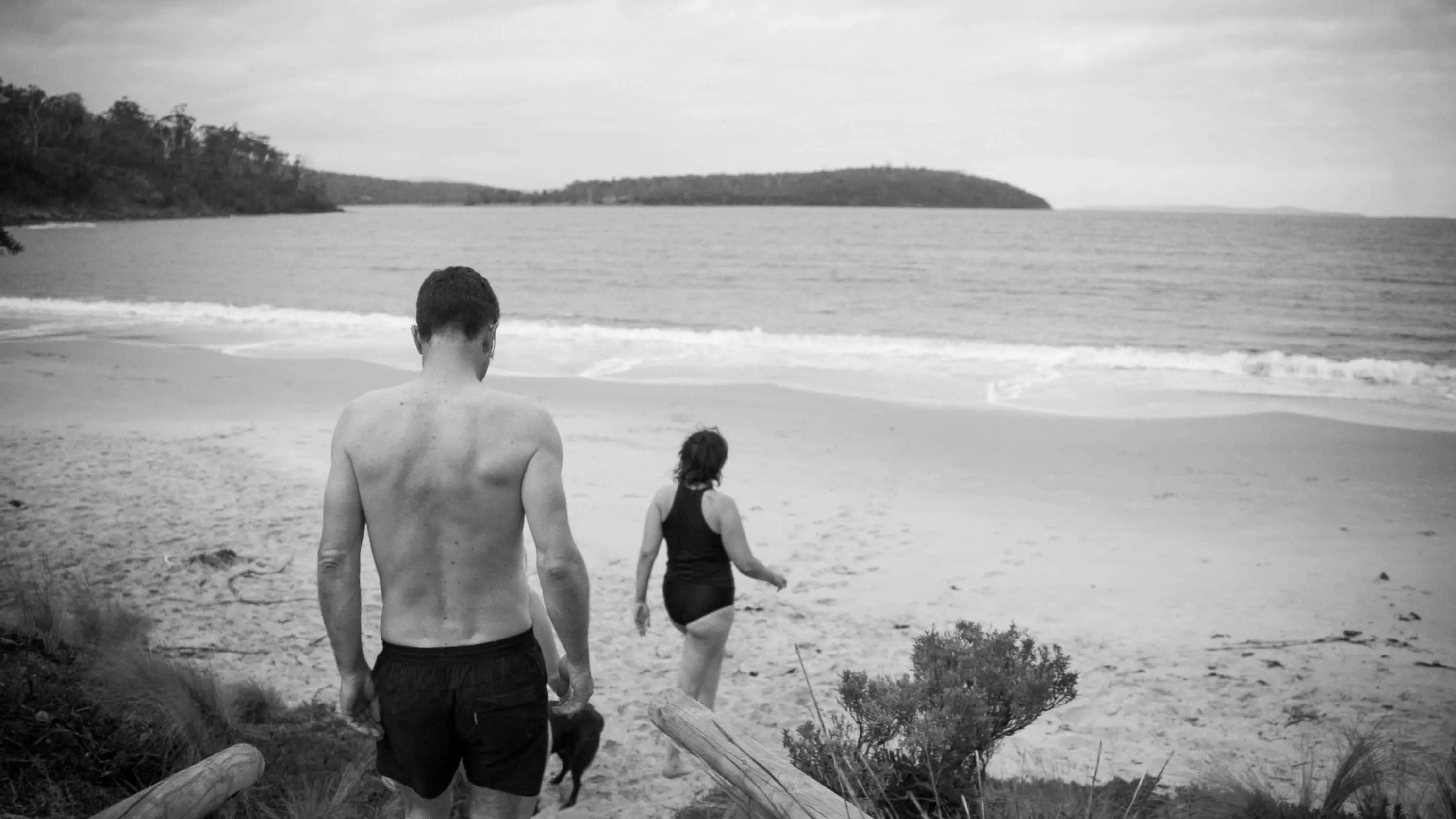 A man and woman wearing swimming gear walk down some steps to the sand on a wide beach towards the water.