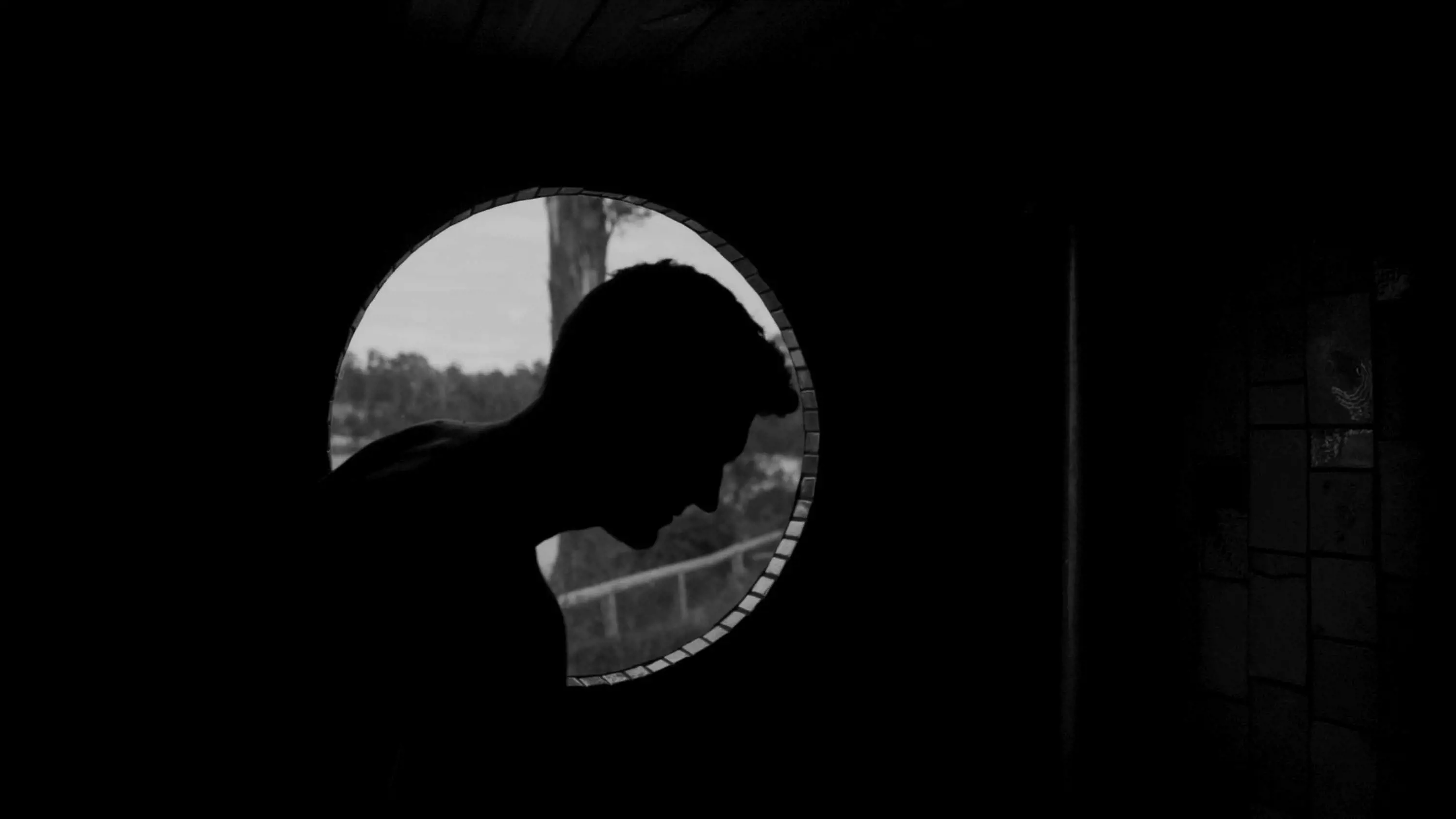 A man's profile silhouette is seen in the backlight from a circular portal window inside a sauna.