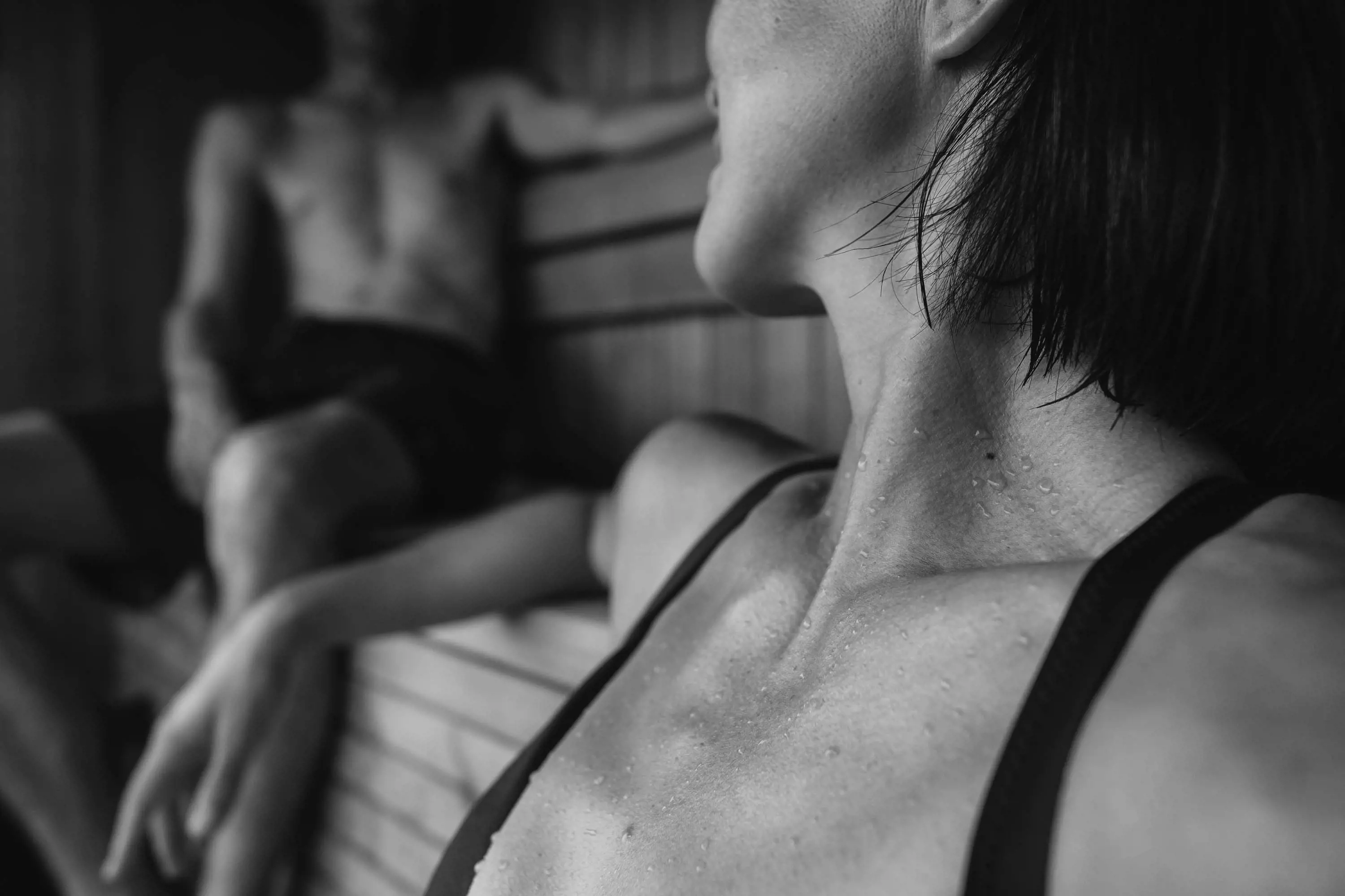 A close up of a woman's face looking over her should to a man who is sitting on a wooden bench in the corner of a large sauna.