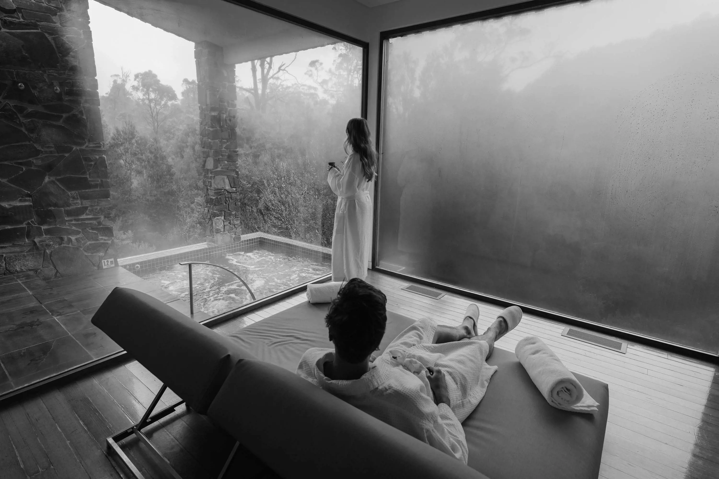 A man wearing a gown made of towelling reclines on a large lounge while a woman wearing a similar gown stands next to a large window looking to an adjacent room with a spa bath and the forest outside.