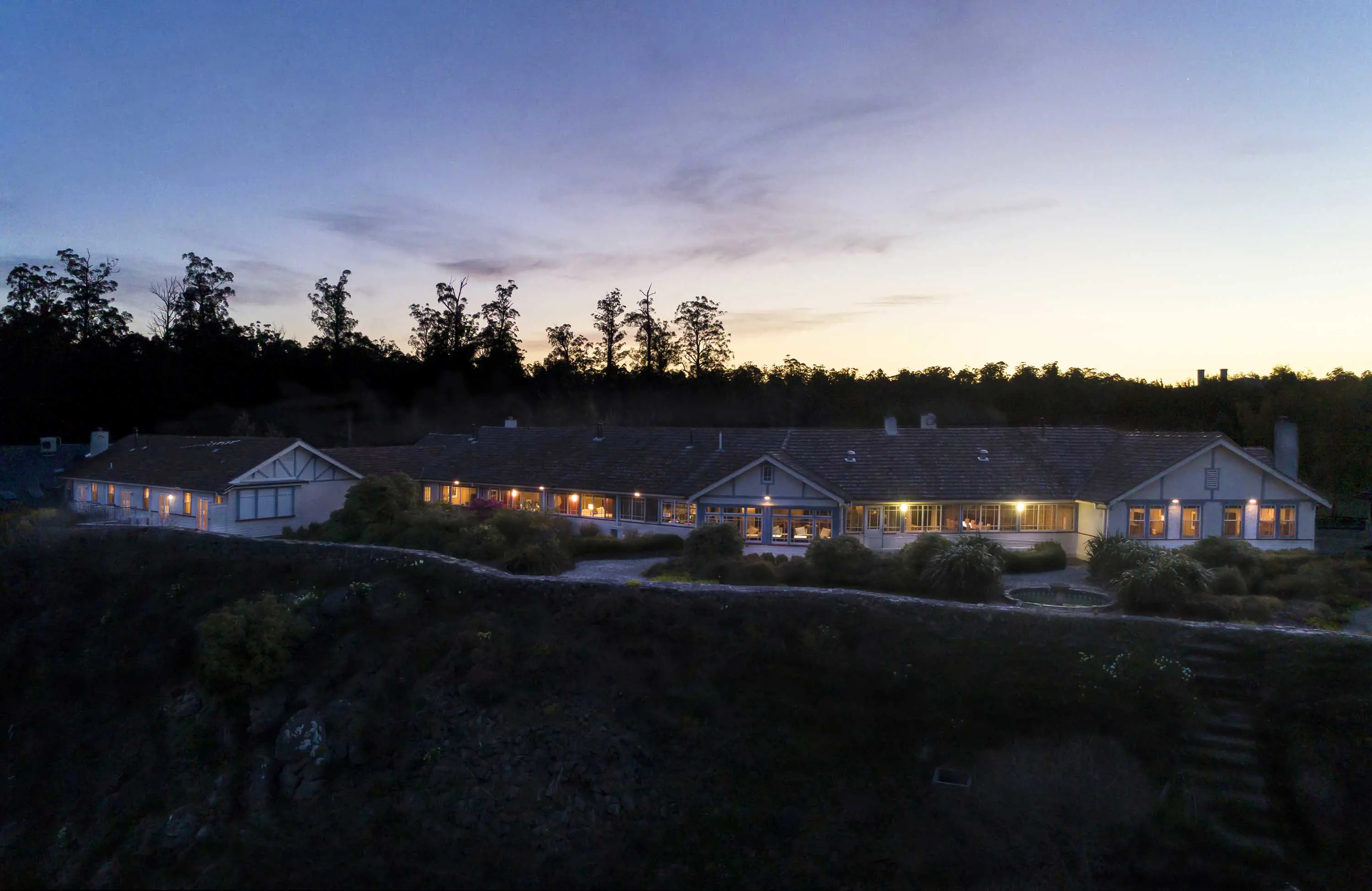 An art deco style building with multiple wings at night time, set on the edge of a hill in thick forest.