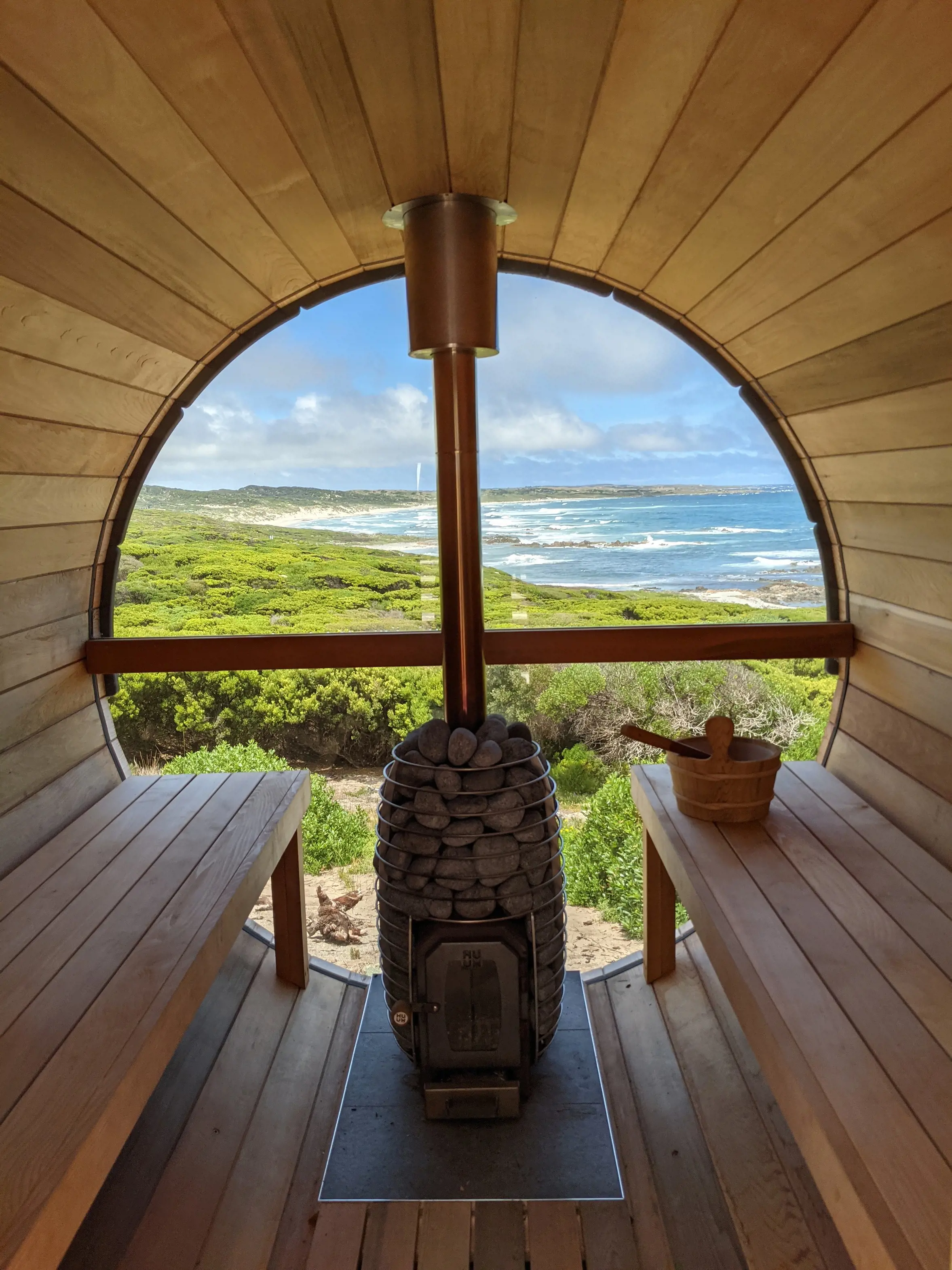 A circular sauna with a central wood fired heater and large circular window looking out over coastal heath and a beach.