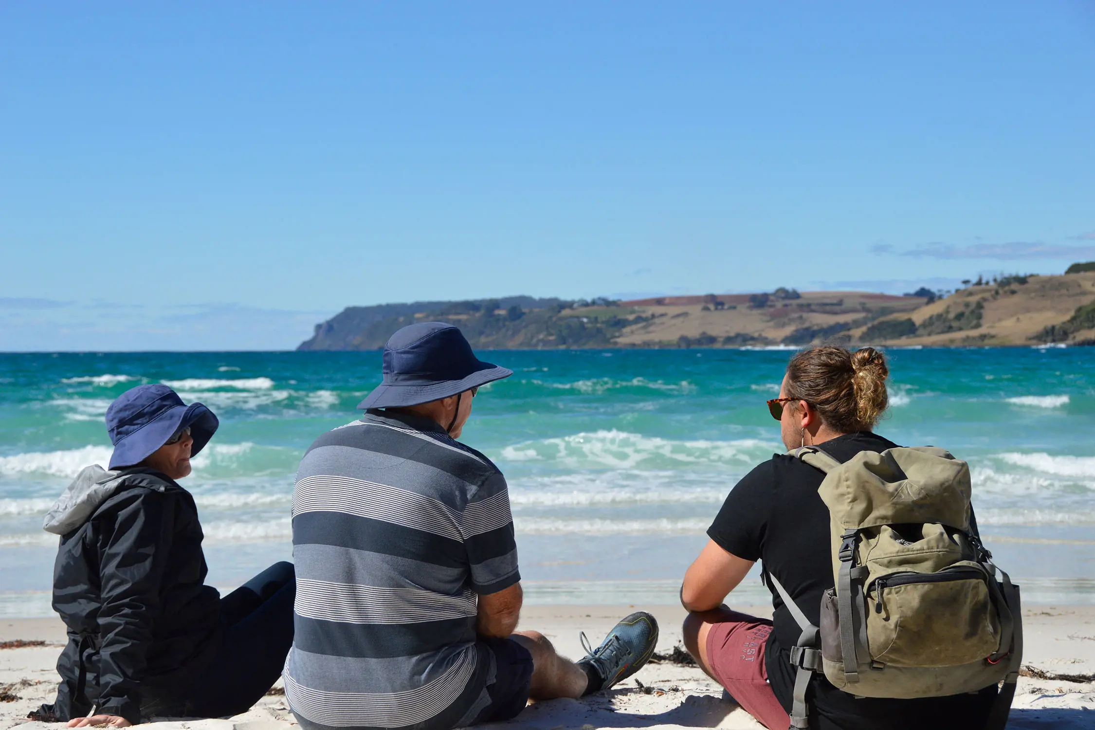 Three people in active outdoors clothing and hats sit on a white sandy beach, facing the aqua blue water.