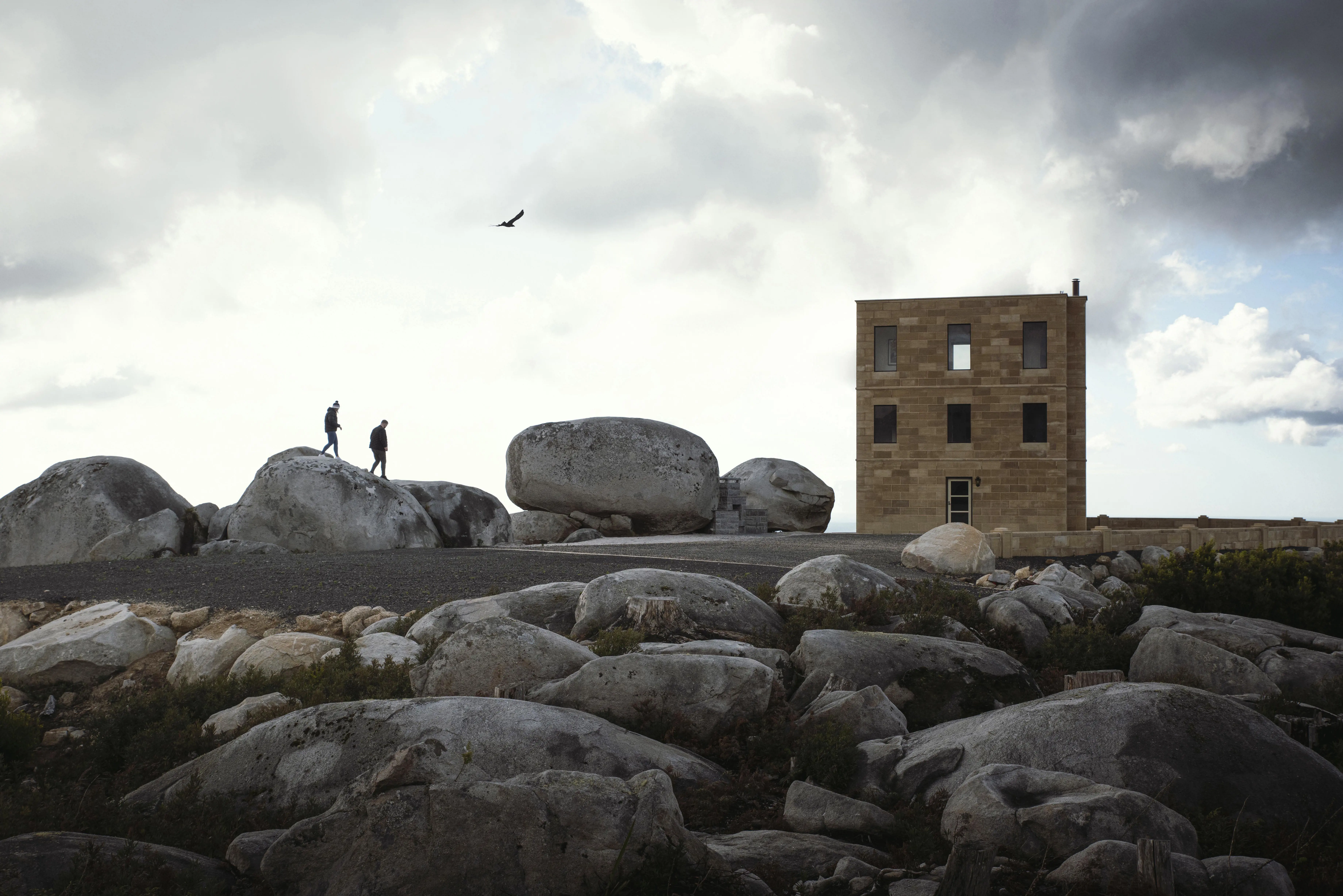 Two people walk along the top of large boulders in front of a modern brick building. 