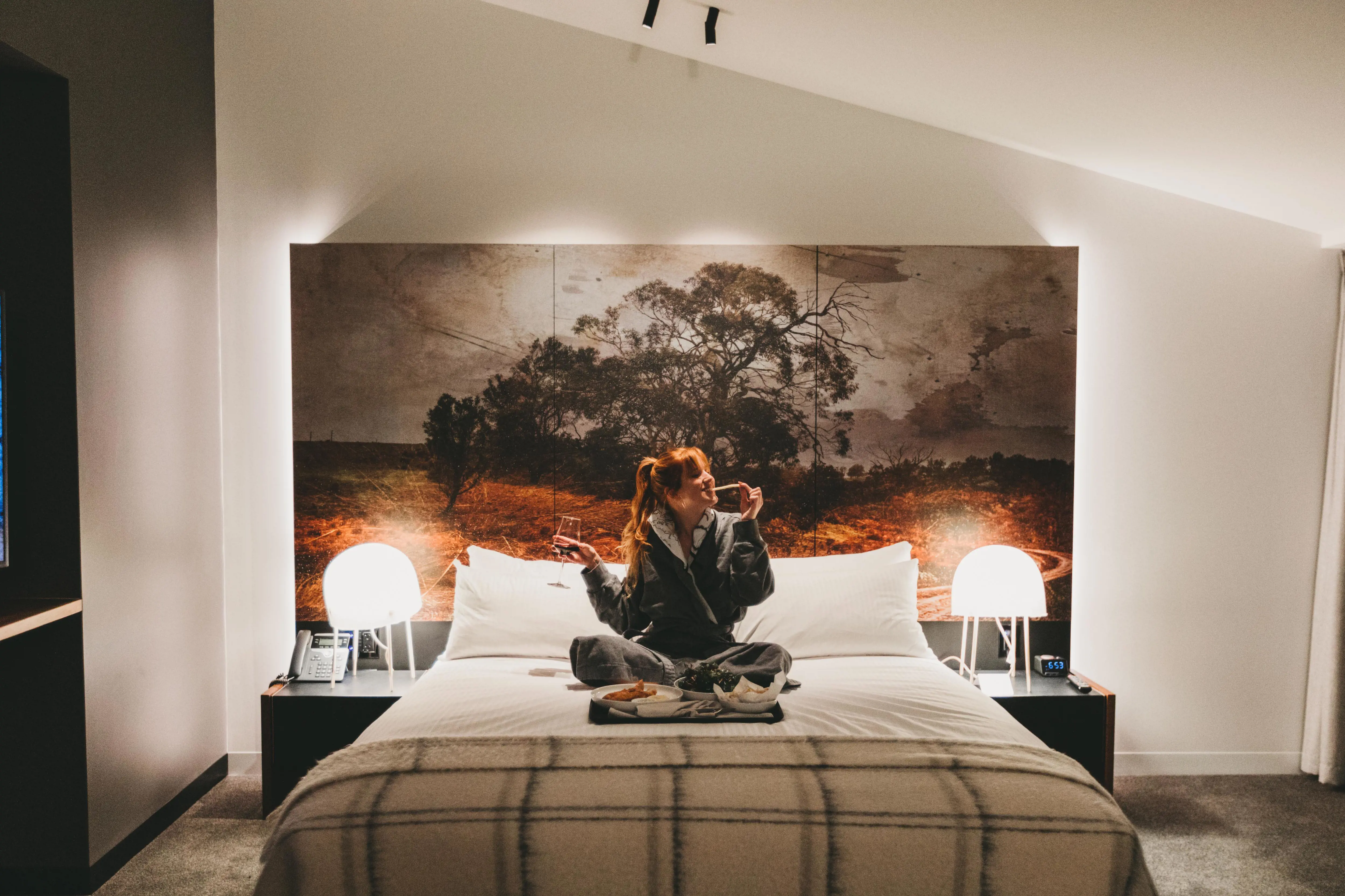 A young woman sits on a large bed in a modern hotel room and enjoys some battered fish and chips. 