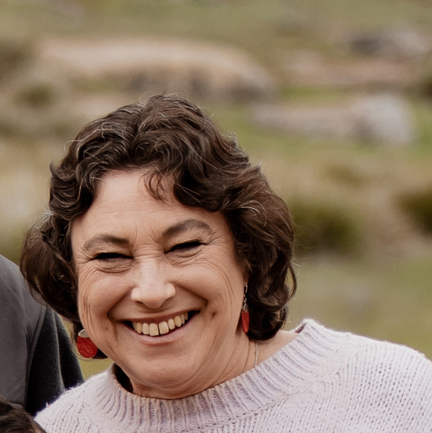 A head-and-shoulders image of a woman with dark brown, bob-length curly hair, wearing a beige jumper. Her face is creased into a huge grin.