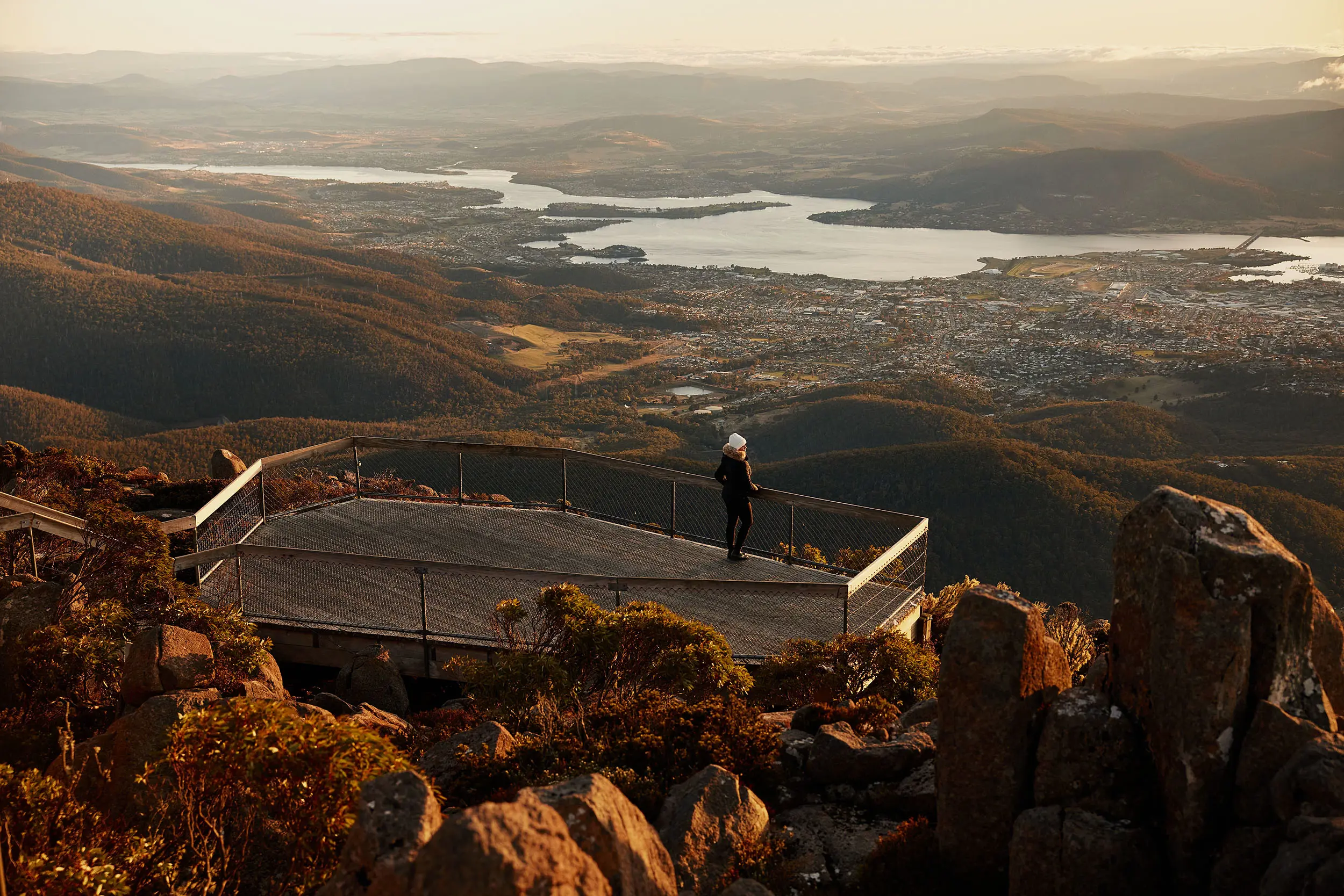 A woman stands at the end of a viewing platform on the side of a mountain overlooking Hobart and the River Derwent at sunrise.