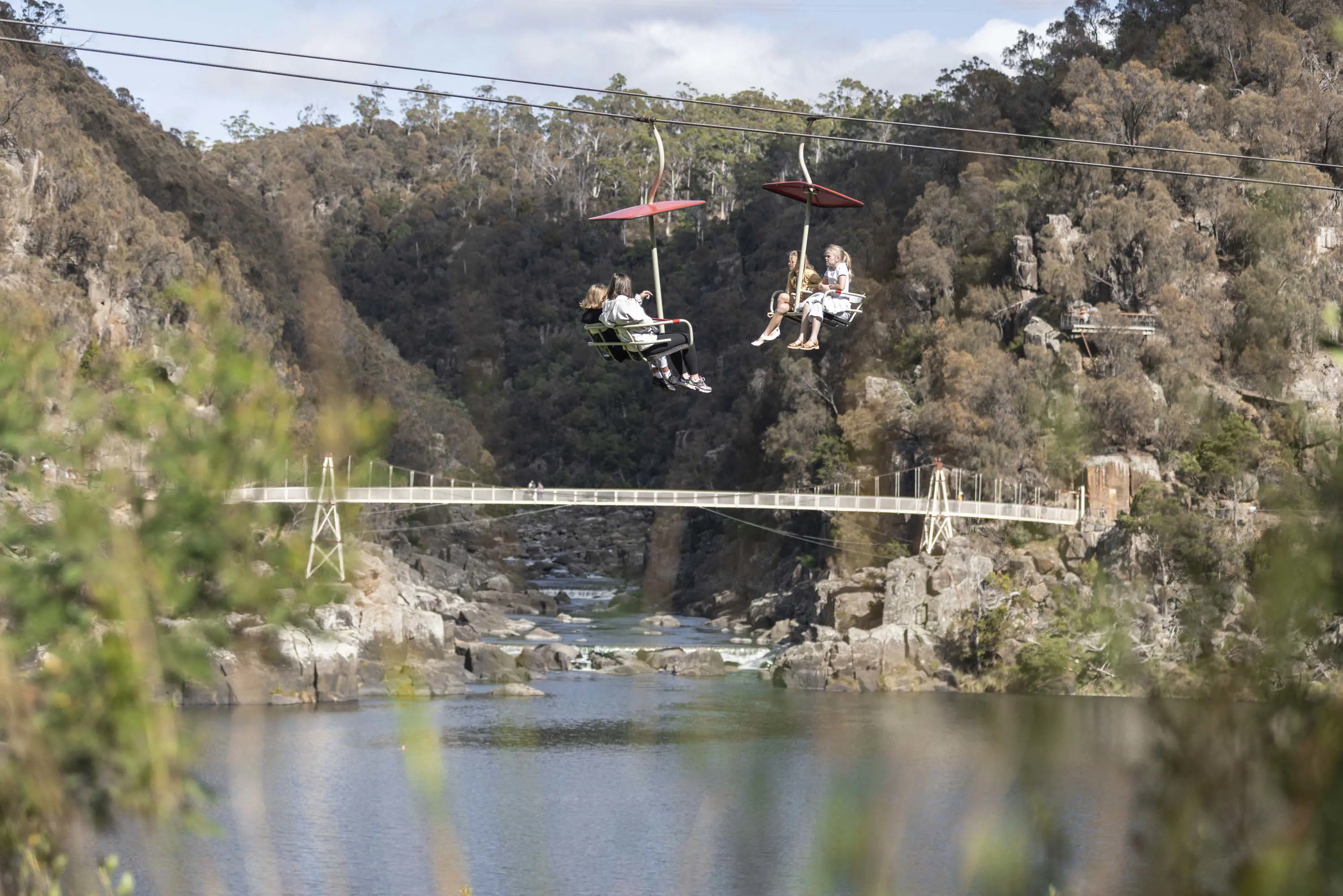 People ride a chairlife attached to cables over water in the Cataract Gorge. A long suspension bridge can be seen in the background.