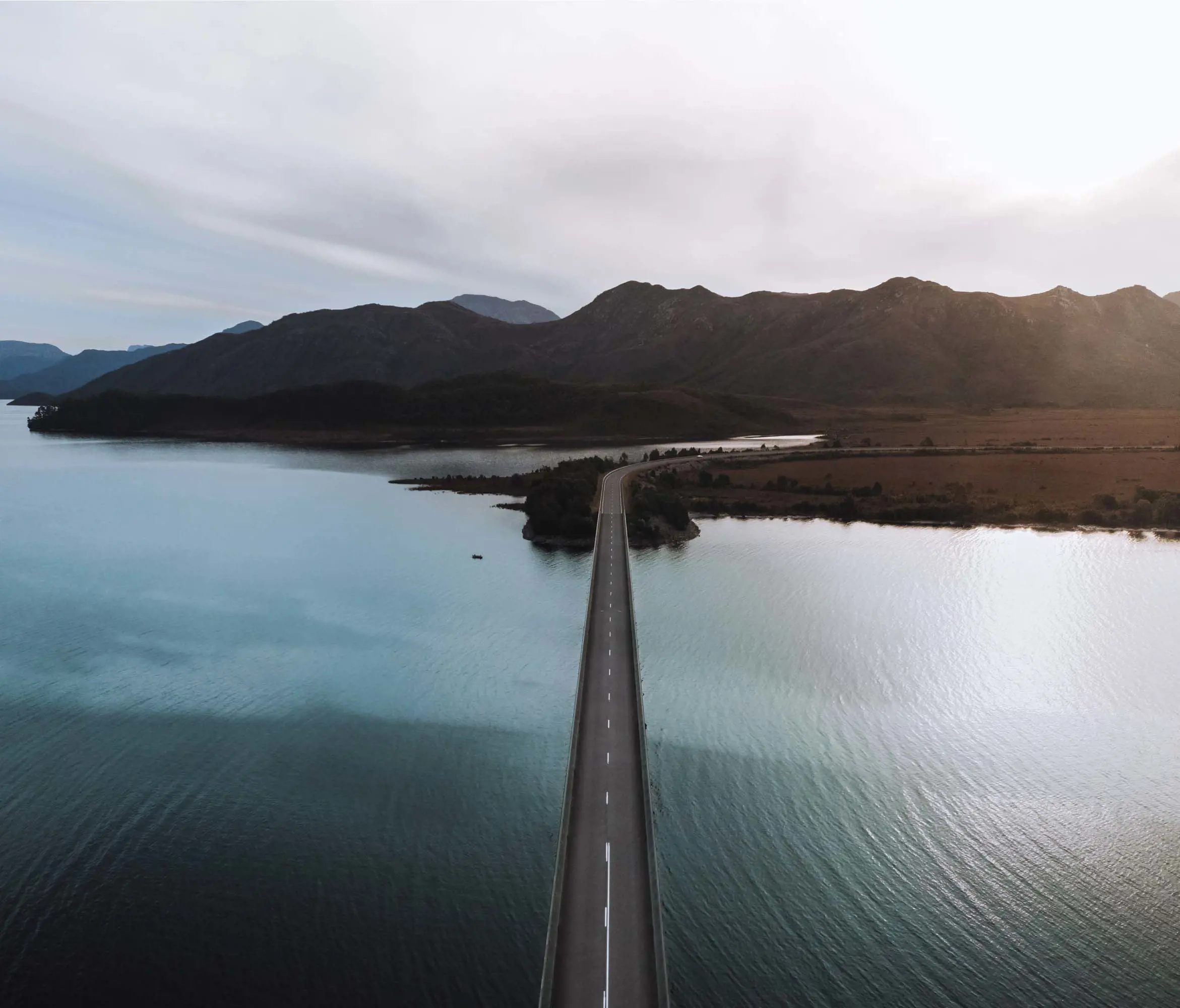 A straight, two-lane bridge extends over a large lake with mountains in the background.