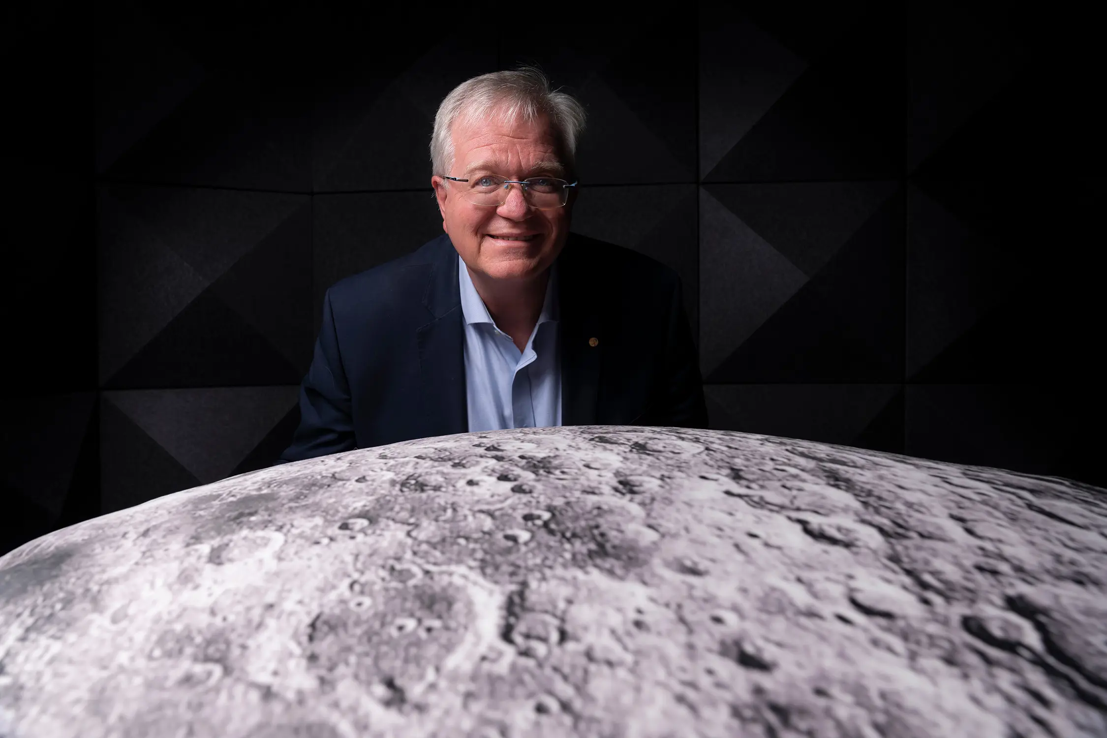 A man in dark suit and glasses stands behind a large spherical model of the moon, positioned against a dark background.