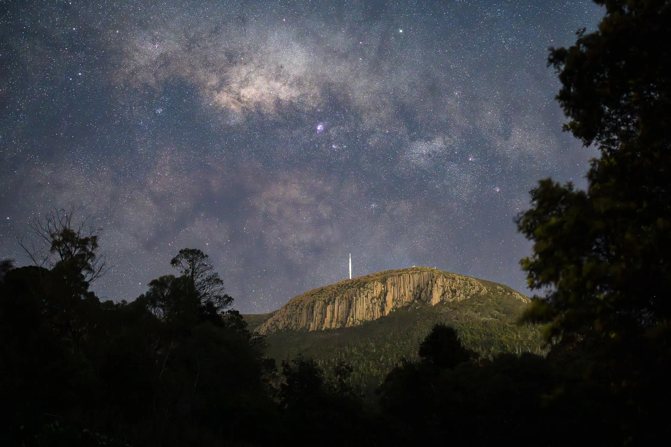 The starry night sky stretches out above a distinctive rocky mountain formation. Silhouetted trees frame the cliff face on both sides.