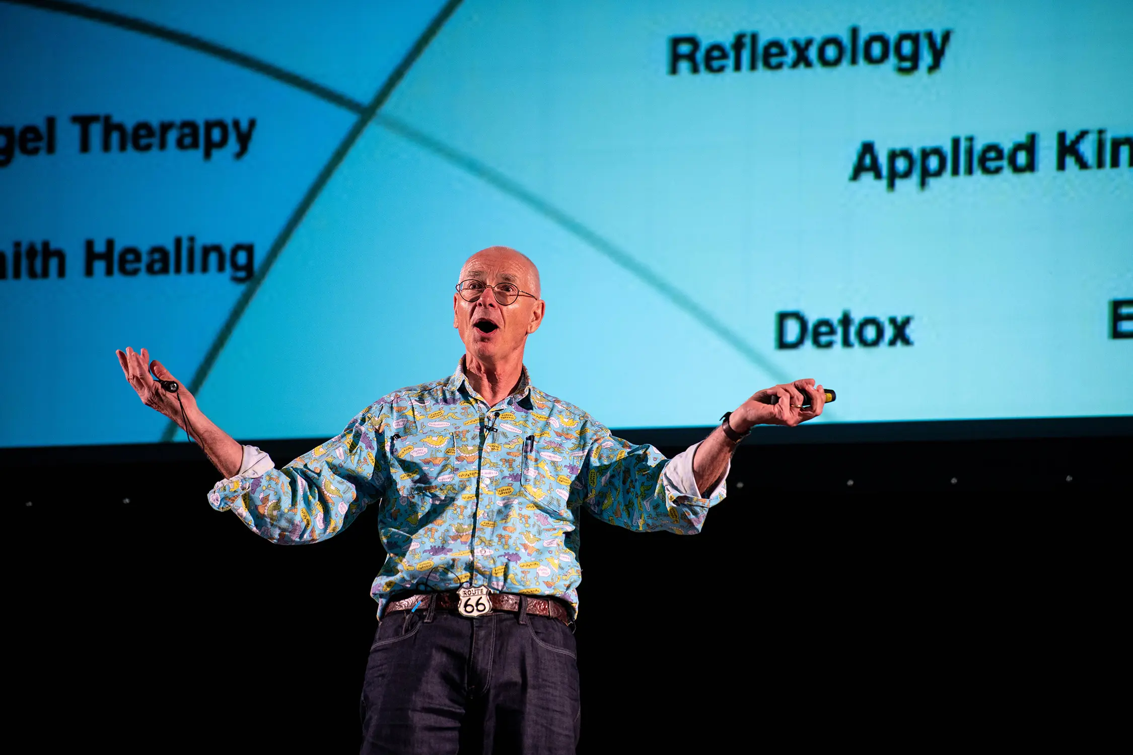 A man in a patterned shirt gestures with both arms raised while speaking on stage. A projection behind him displays text including "Reflexology" and "Detox."