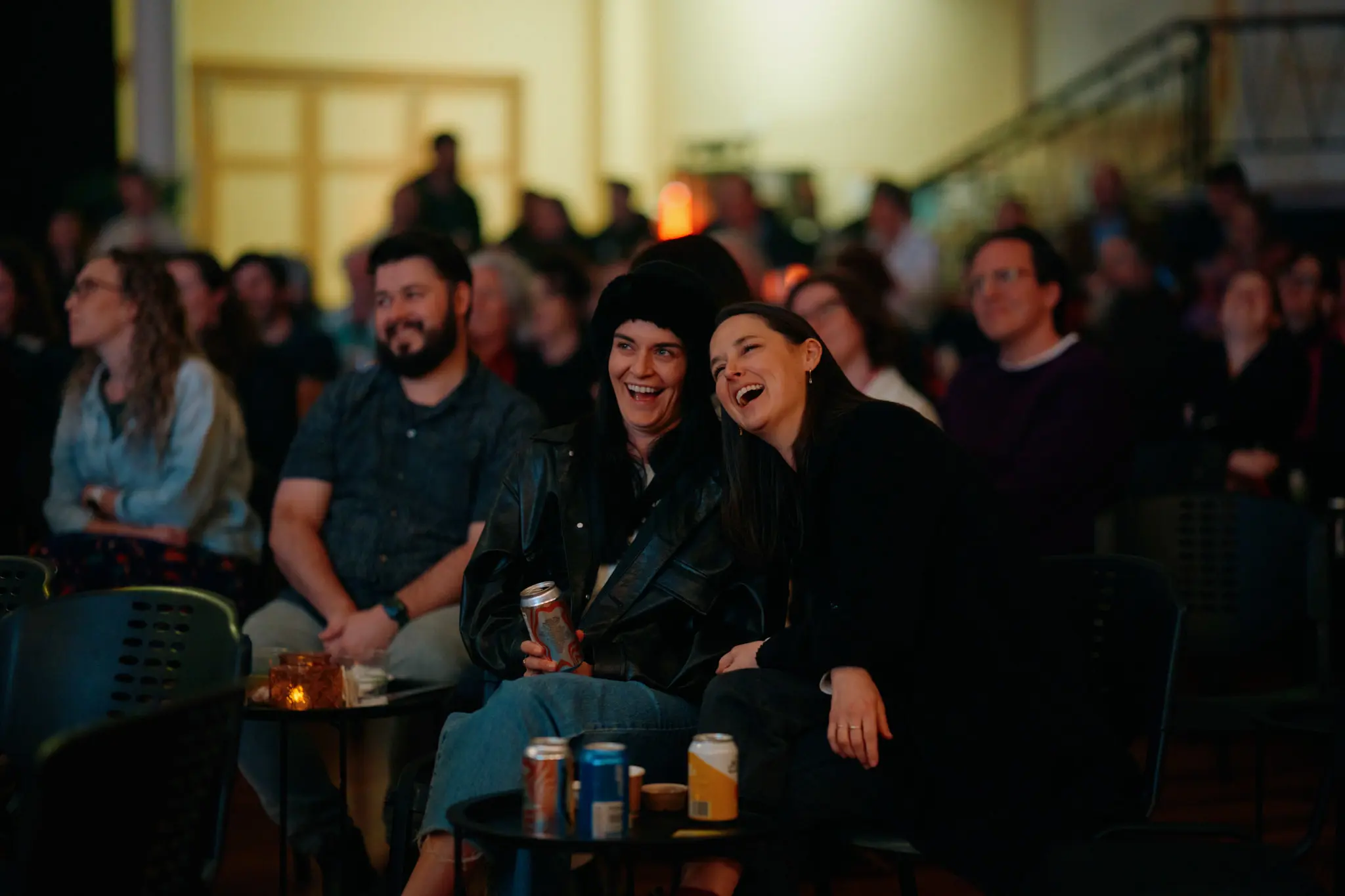 Audience members laugh while seated at small tables with drinks in a dimly lit indoor venue.