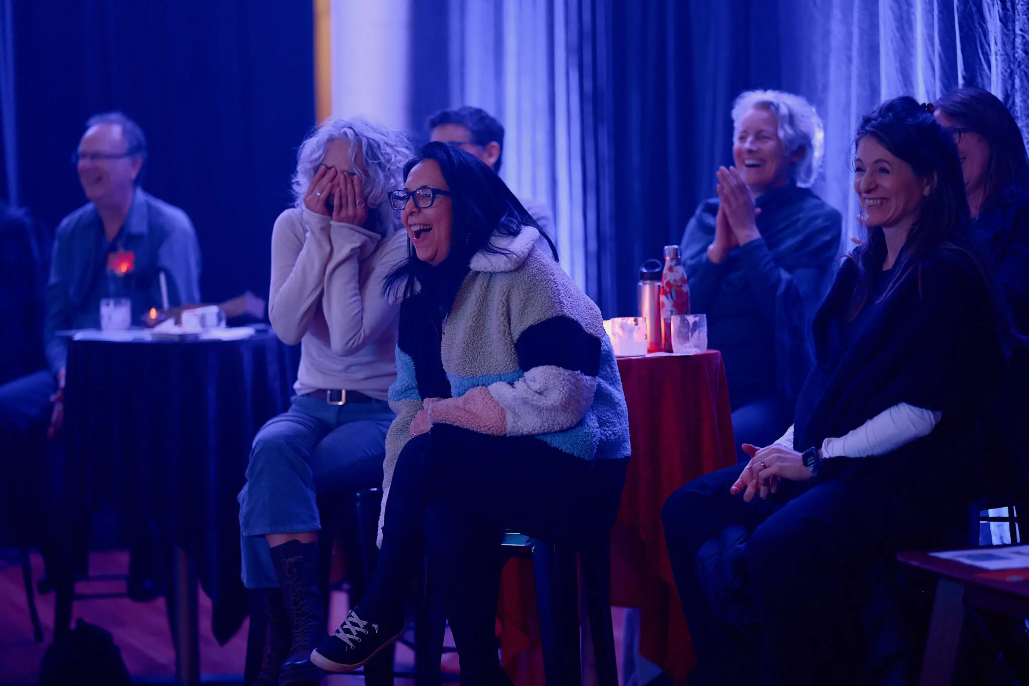 Audience members sit on bar stools at tall tables under blue and purple stage lighting, laughing and clapping.