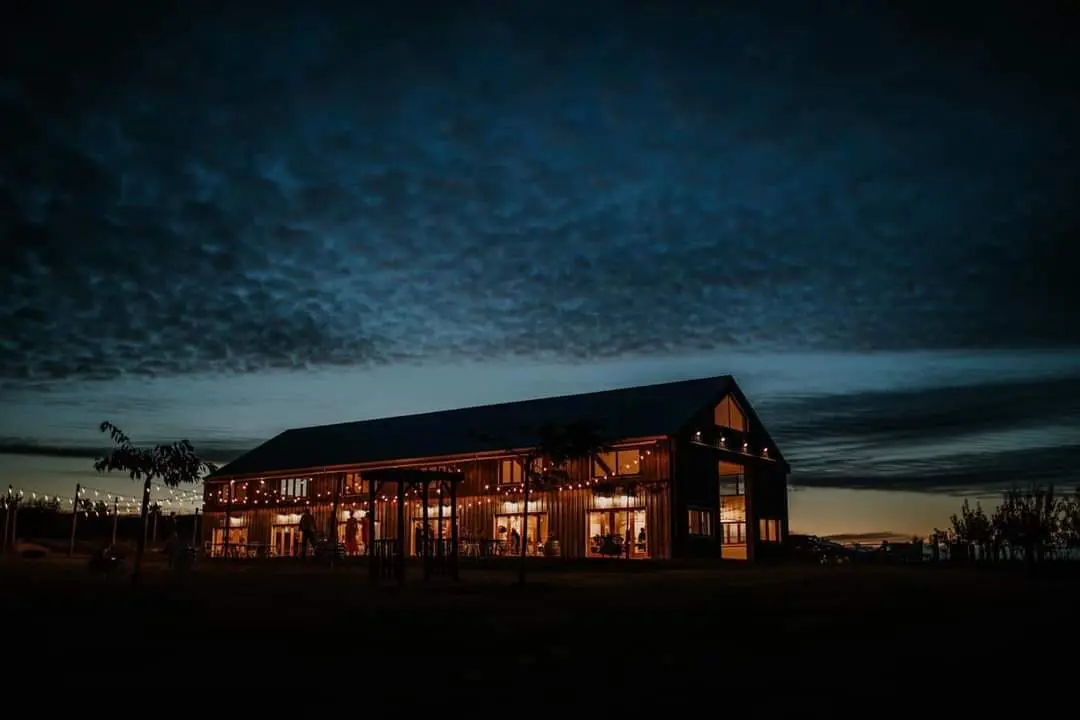 Illuminated rustic building stands against a twilight sky with dramatic cloud formations. The structure features multiple windows glowing with warm interior light.