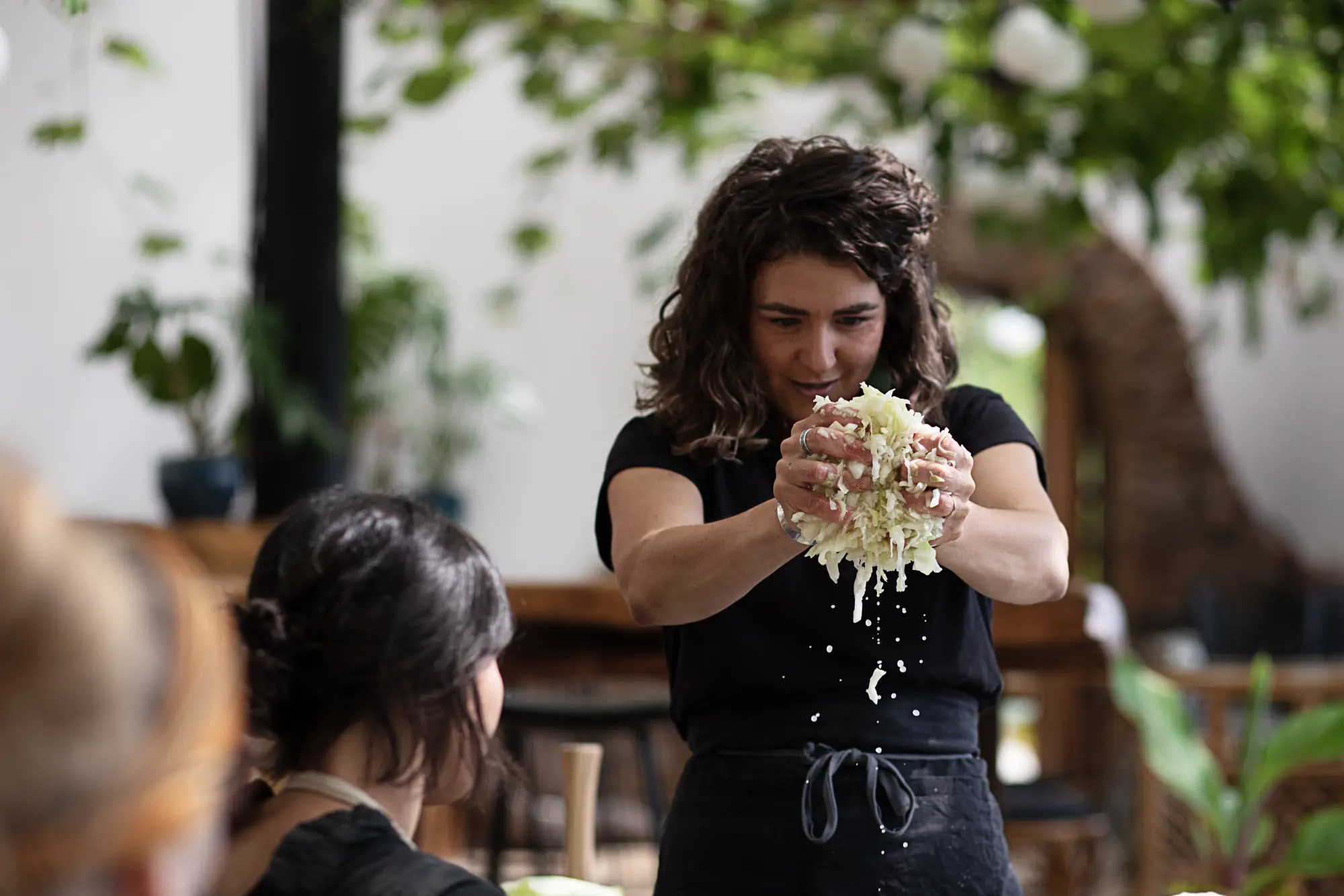 A woman in a dark apron squeezes liquid from a large handful of white cabbage. An audience member watches from the foreground in a bright interior space.