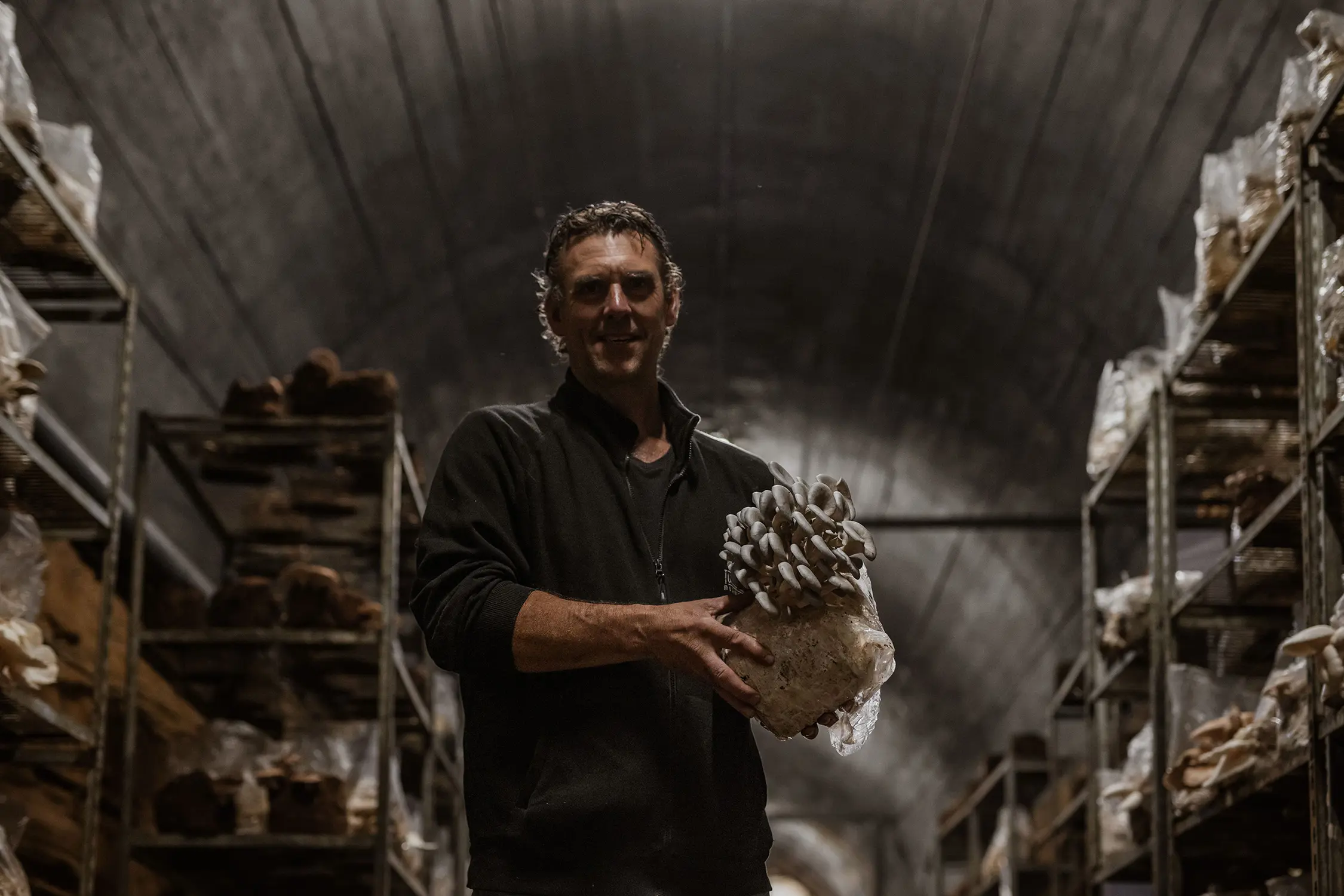A man holds a large mushroom cluster while standing in a dimly lit tunnel. Shelving units with growing materials line both sides.