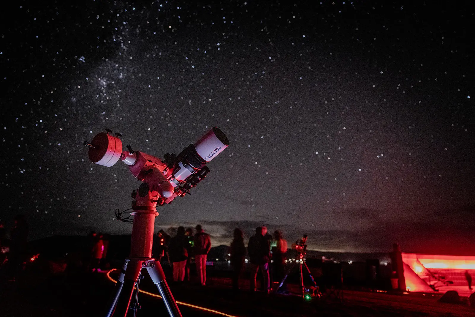 A telescope on a tripod points toward a starry night sky. Silhouetted people gather around the equipment with a glowing screen visible to the right.