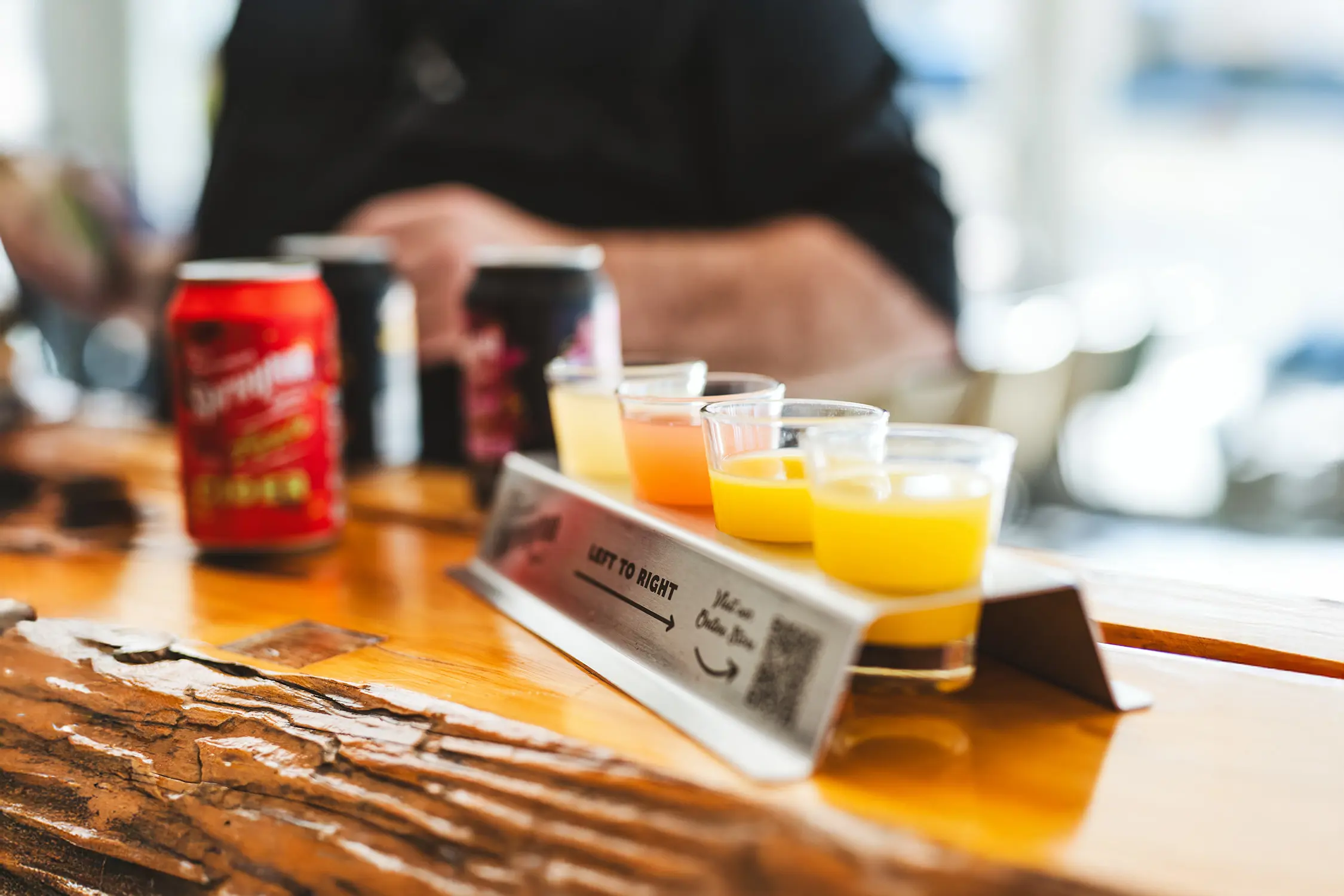 A tasting paddle of four cider glasses, in a stainless steel holder, sit atop a wooden bar. Cans and people drinking are out-of-focus in the background.
