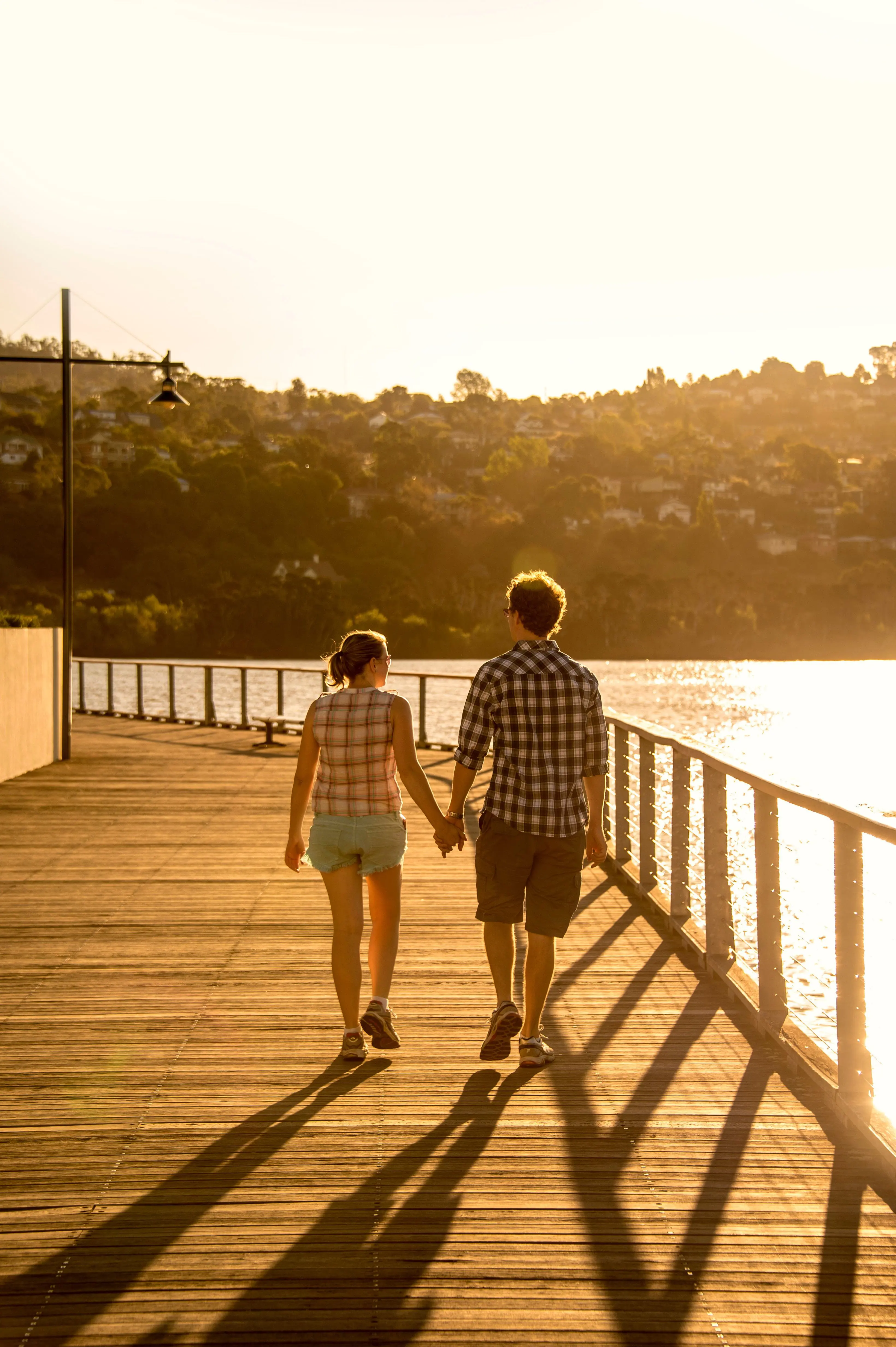A couple in plaid shirts and shorts holds hands while walking along a wooden boardwalk beside the sea with a bushy hill in the background, at golden hour.