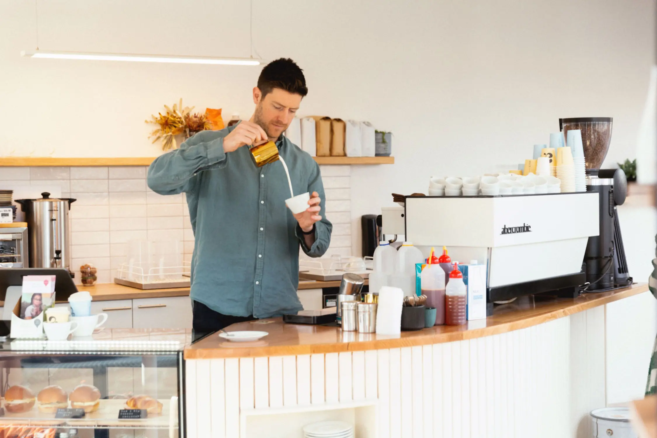 The interior of a coffee shop shows a staff member pouring coffee behind a counter with white cabinetry and shelving.