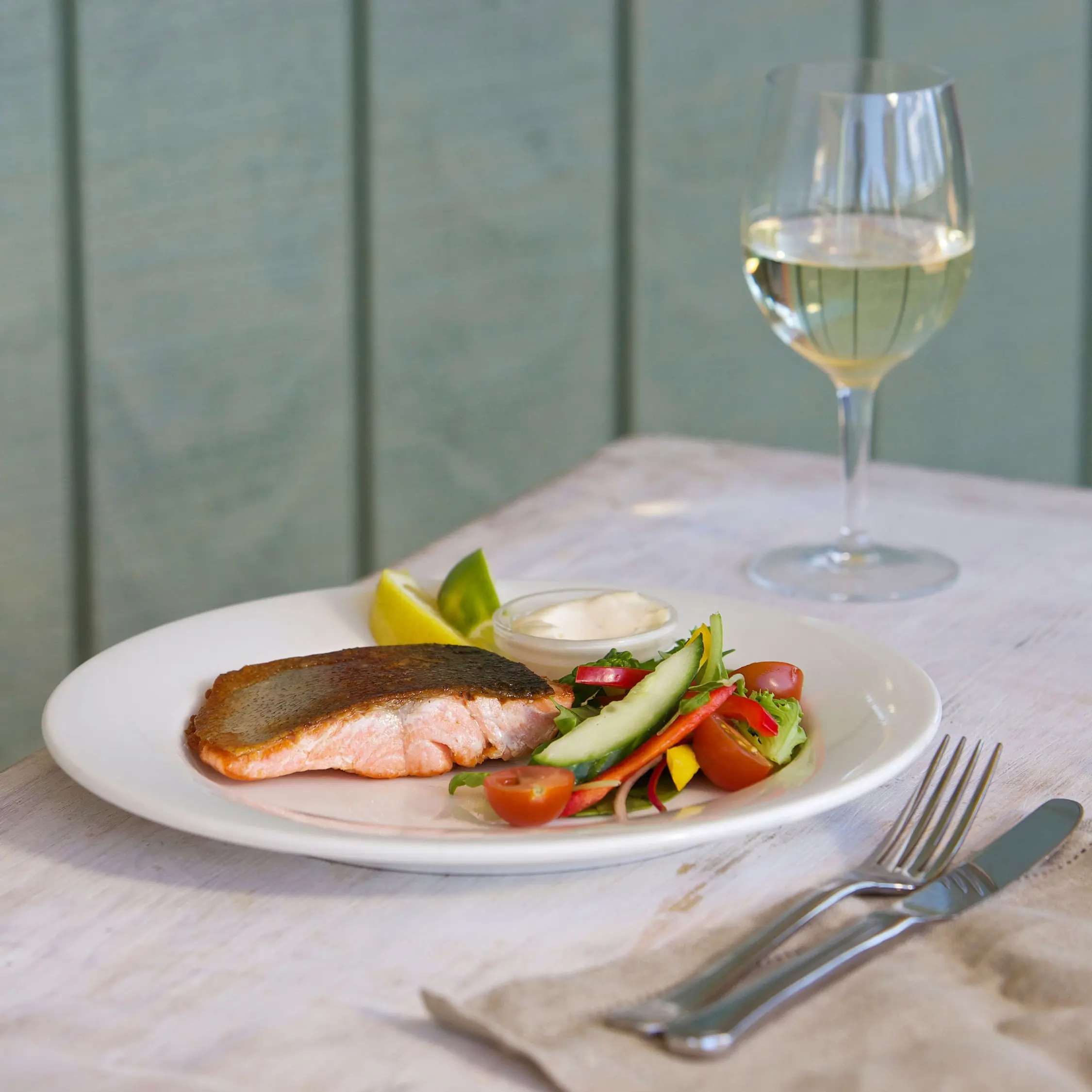 A white plate holds cooked salmon fillet with roasted vegetables beside a glass of white wine.
