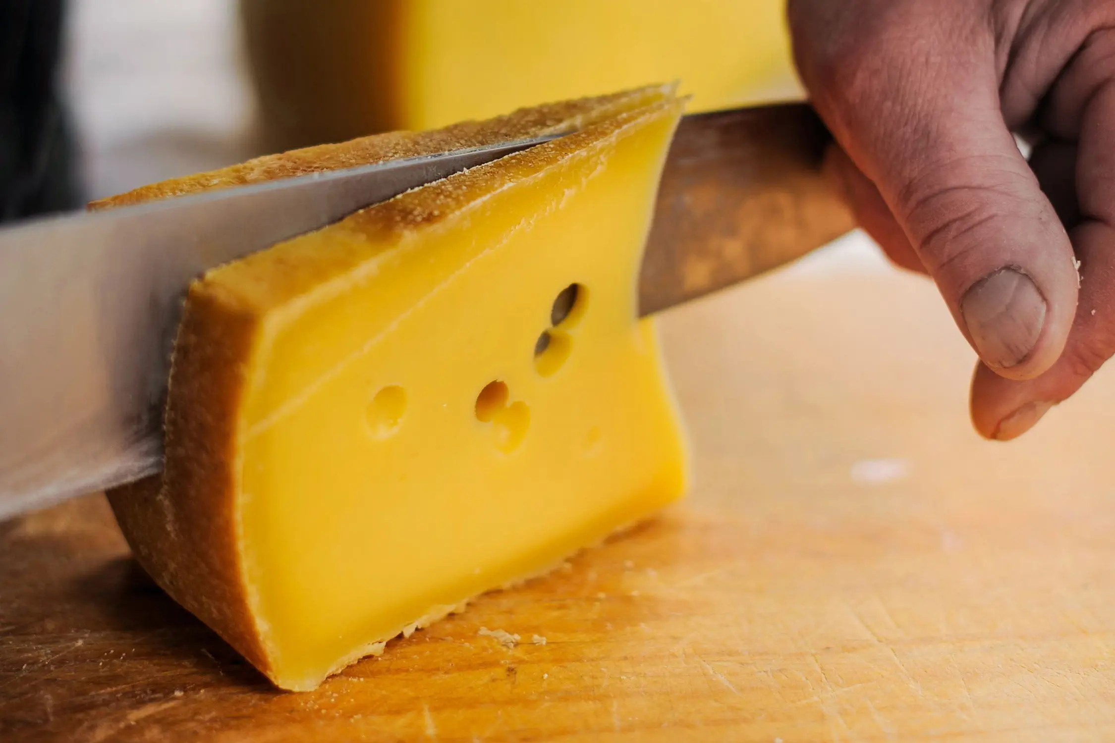 A hand uses a knife to slice bright yellow cheese on a wooden cutting board.