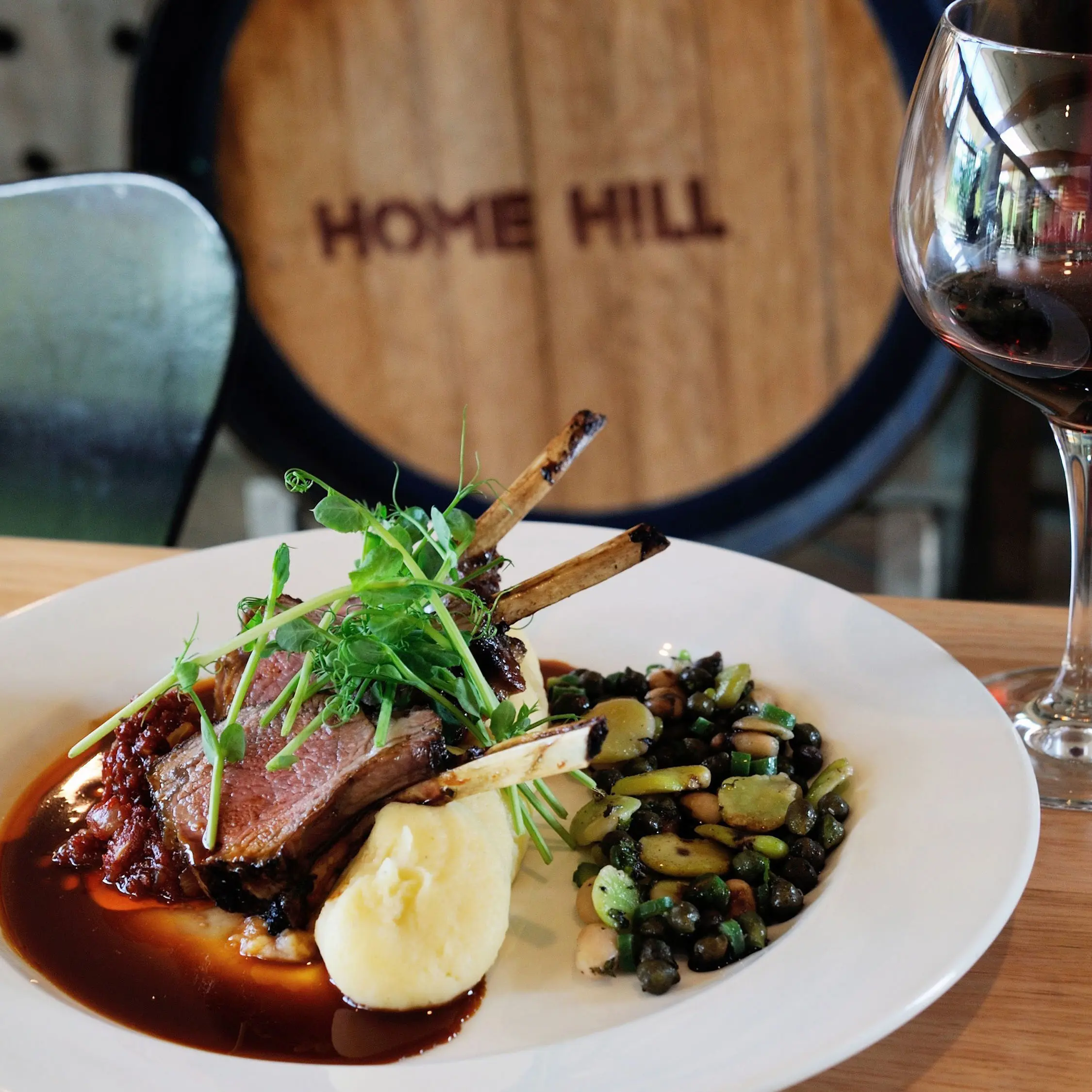 A white plate holds a cooked roast with smooth mash potato, red wine sauce and leafy garnish. A wine barrel in the background is printed with "Home Hill" on it.