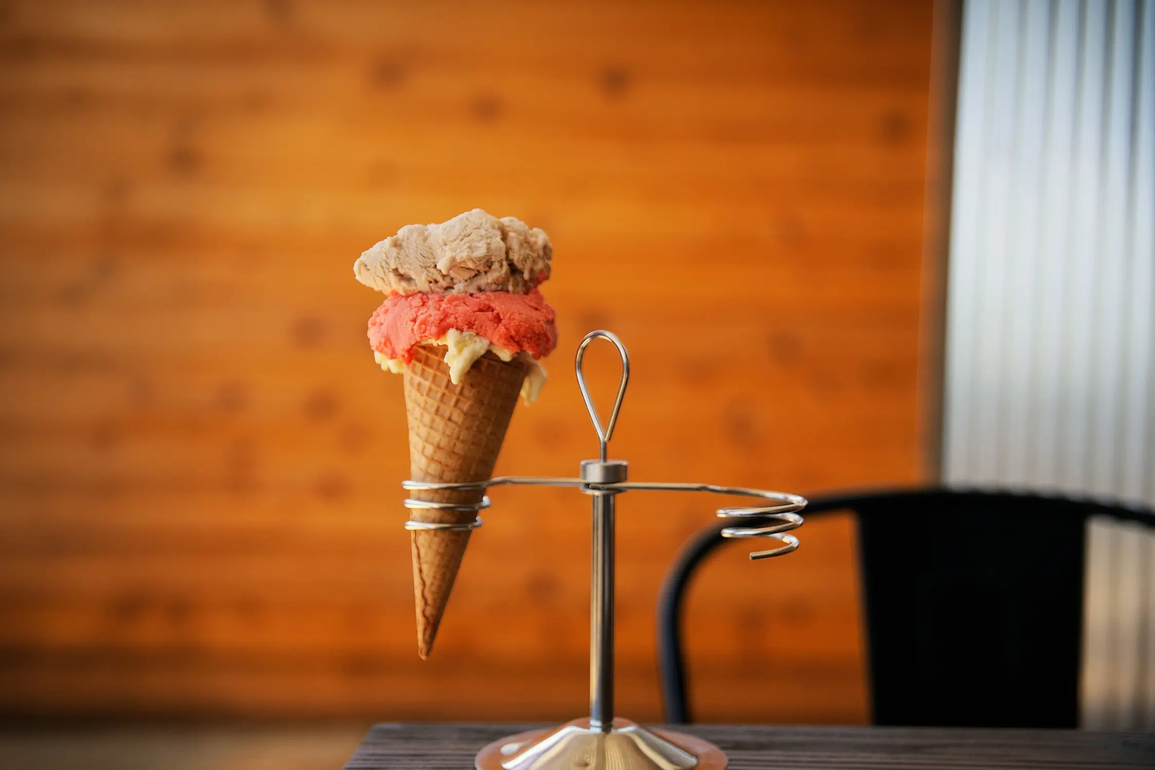 A two-scoop ice cream cone with pink and chocolatey flavours sits in a metal stand against a timber-clad wall.