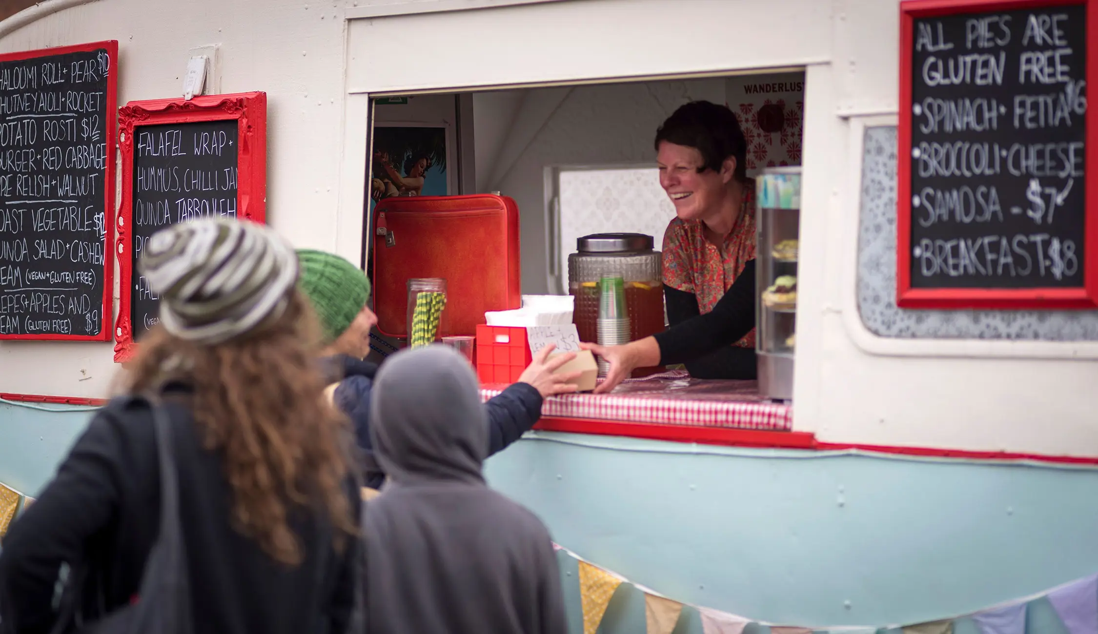 People queue at the serving window of a blue and white vintage caravan food truck, with chalkboard menus displaying food options.