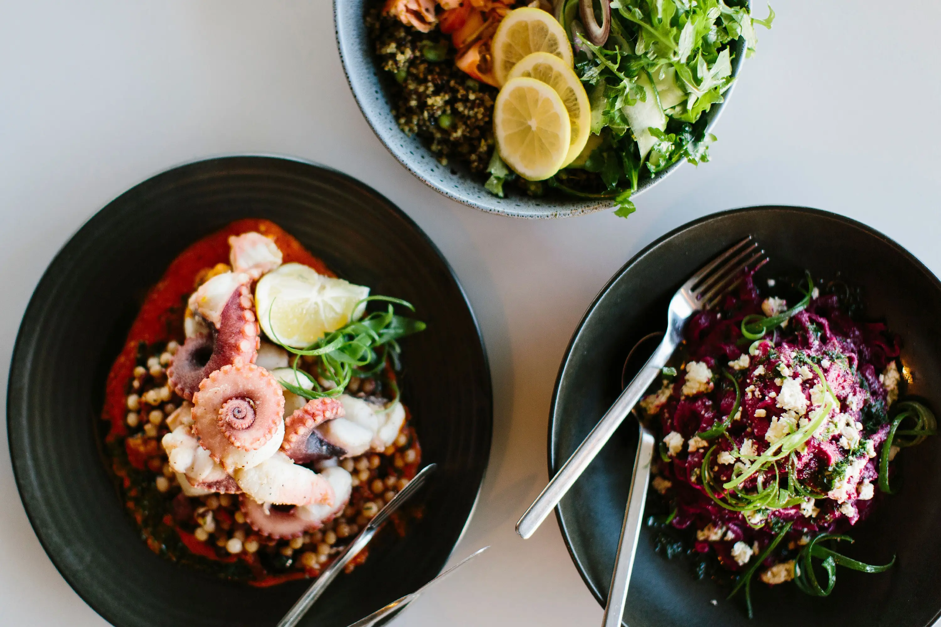 Three black plates display artfully colourful meals, including one with lemon slices and fresh greens, another with beetroot and feta, and a third with octopus on a salad bed.