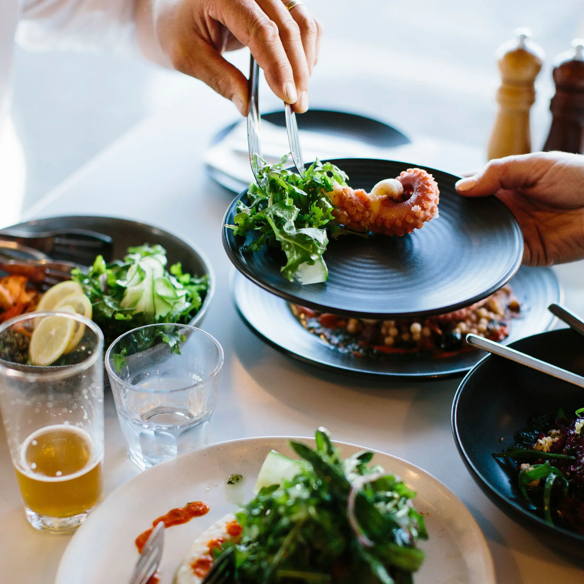 People dish up salad greens and fried octopus from share plates at a table laid with drinks and a white tablecloth.