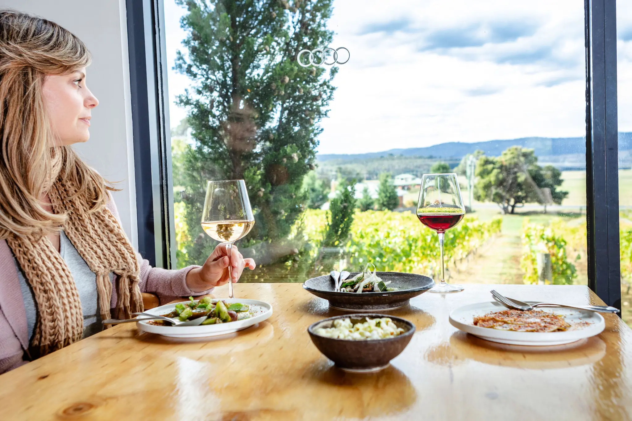 A woman holds a glass of white wine at a wooden table, looking toward vineyard views through large windows.