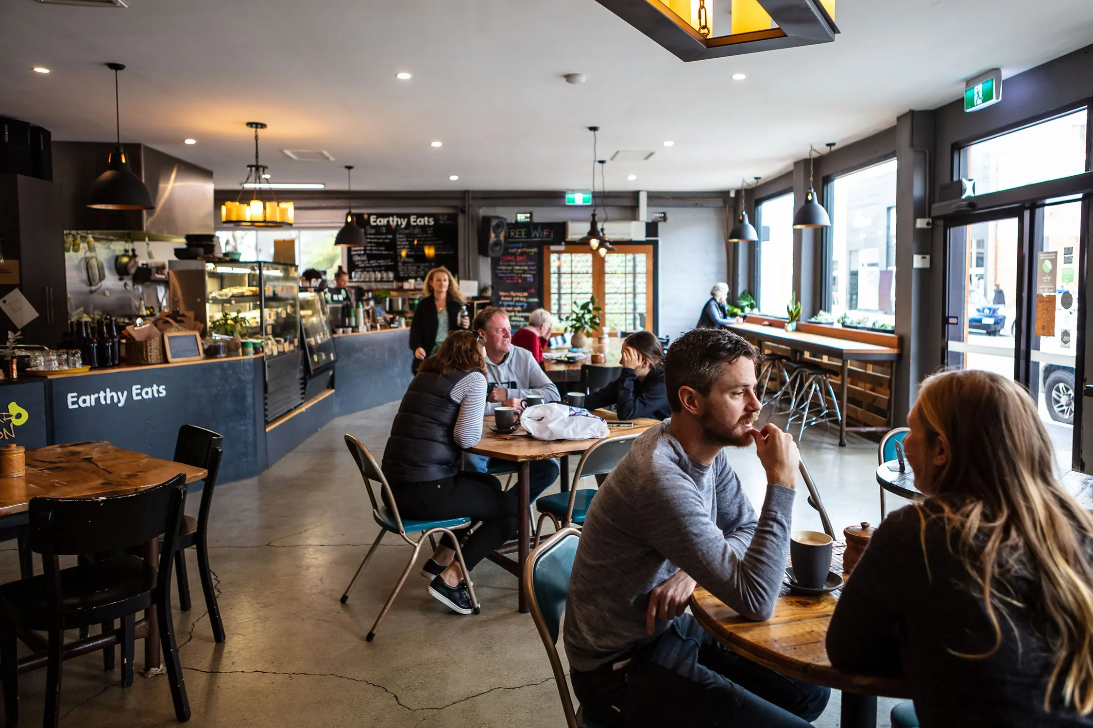 The interior of a cafe shows customers seated at wooden tables with pendant lighting and a service counter.