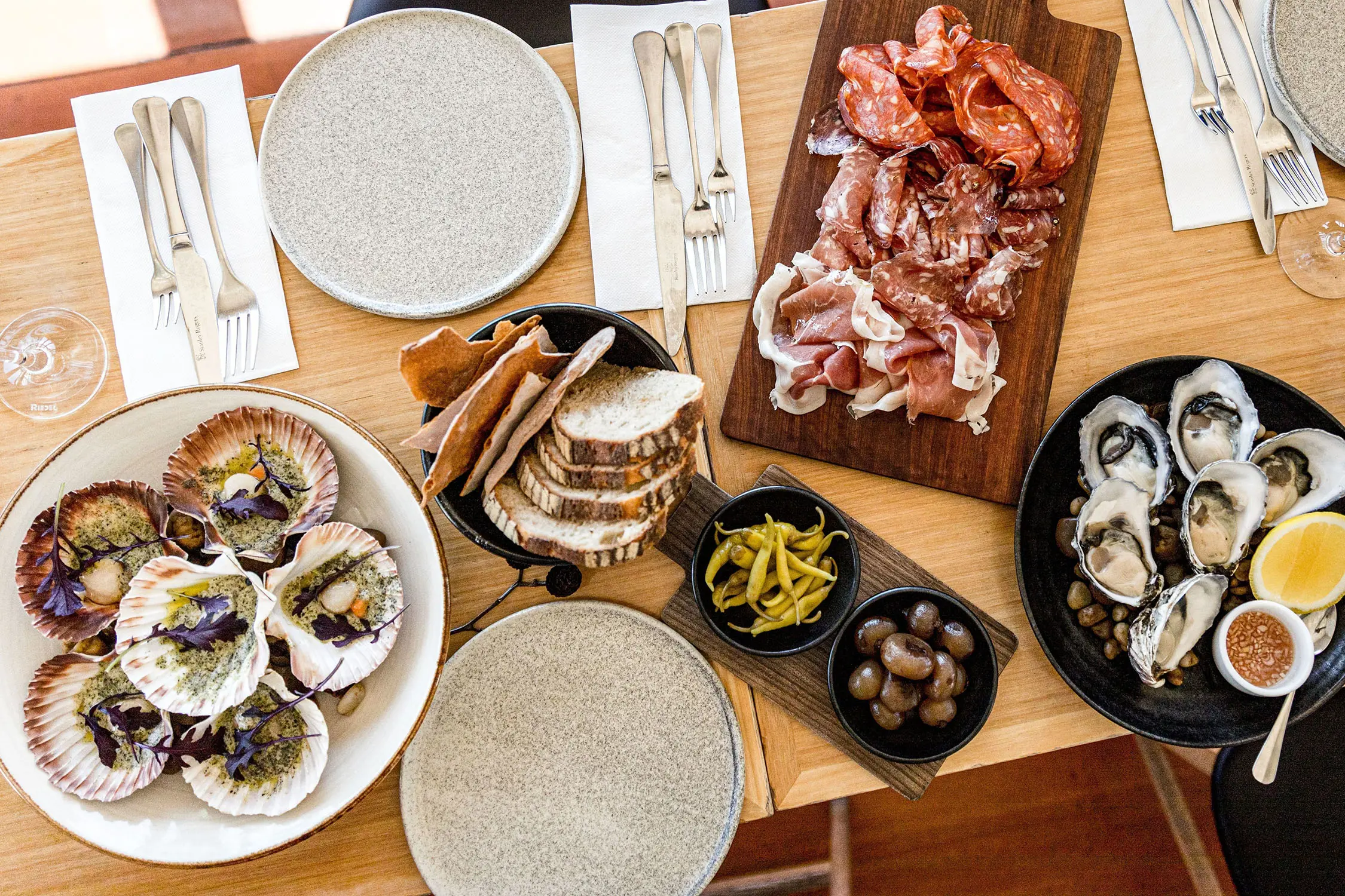 An overhead view shows multiple dishes on a wooden table including oysters, scallops and cured meats.