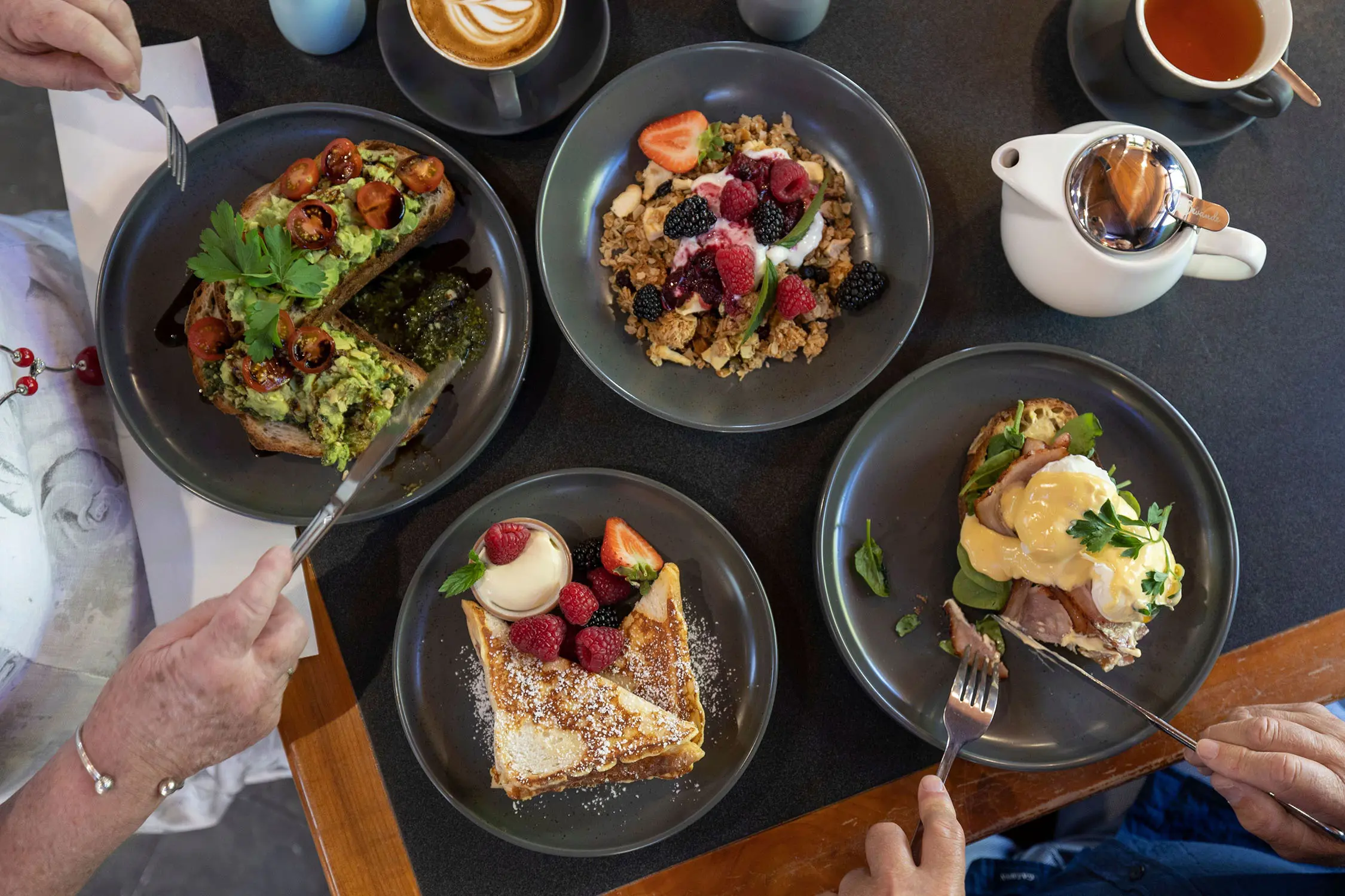 Multiple dark plates display colourful dishes with berries, greens and various toppings viewed from above.